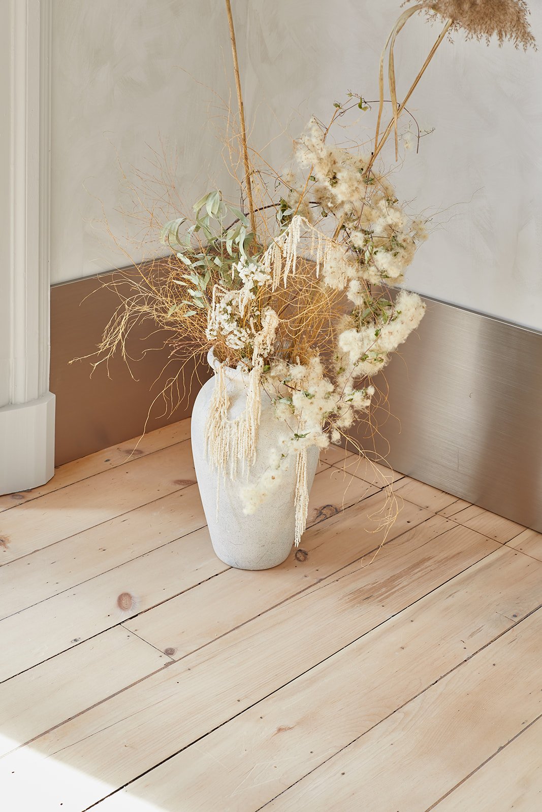 A white vase with dried and preserved flowers and foliage, including pampas grass and eucalyptus, placed on a light wooden floor against a wall.