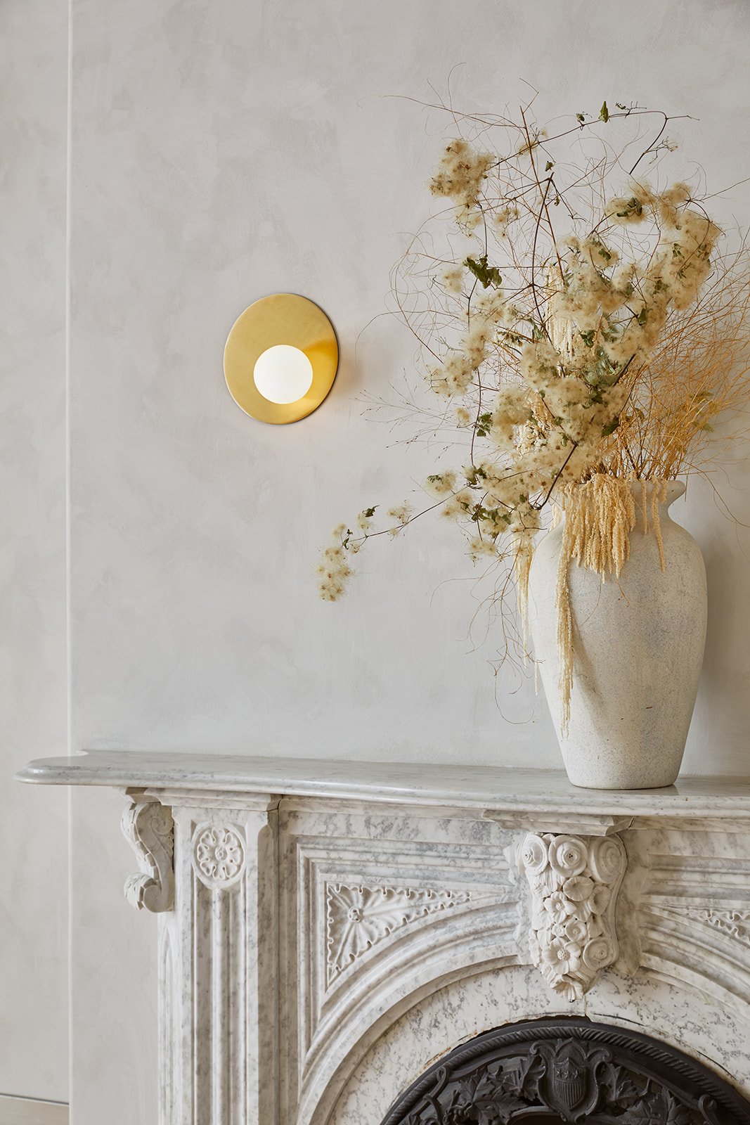 A beige ceramic vase with dried flowers and grasses on a white marble mantelpiece, with a minimalist round wall light on a light-colored wall.