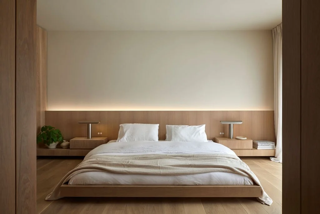 A tranquil, minimalist primary bedroom featuring a low-profile wood platform bed, wide-plank hardwood floors, and a custom integrated oak headboard with soft ambient backlighting.
