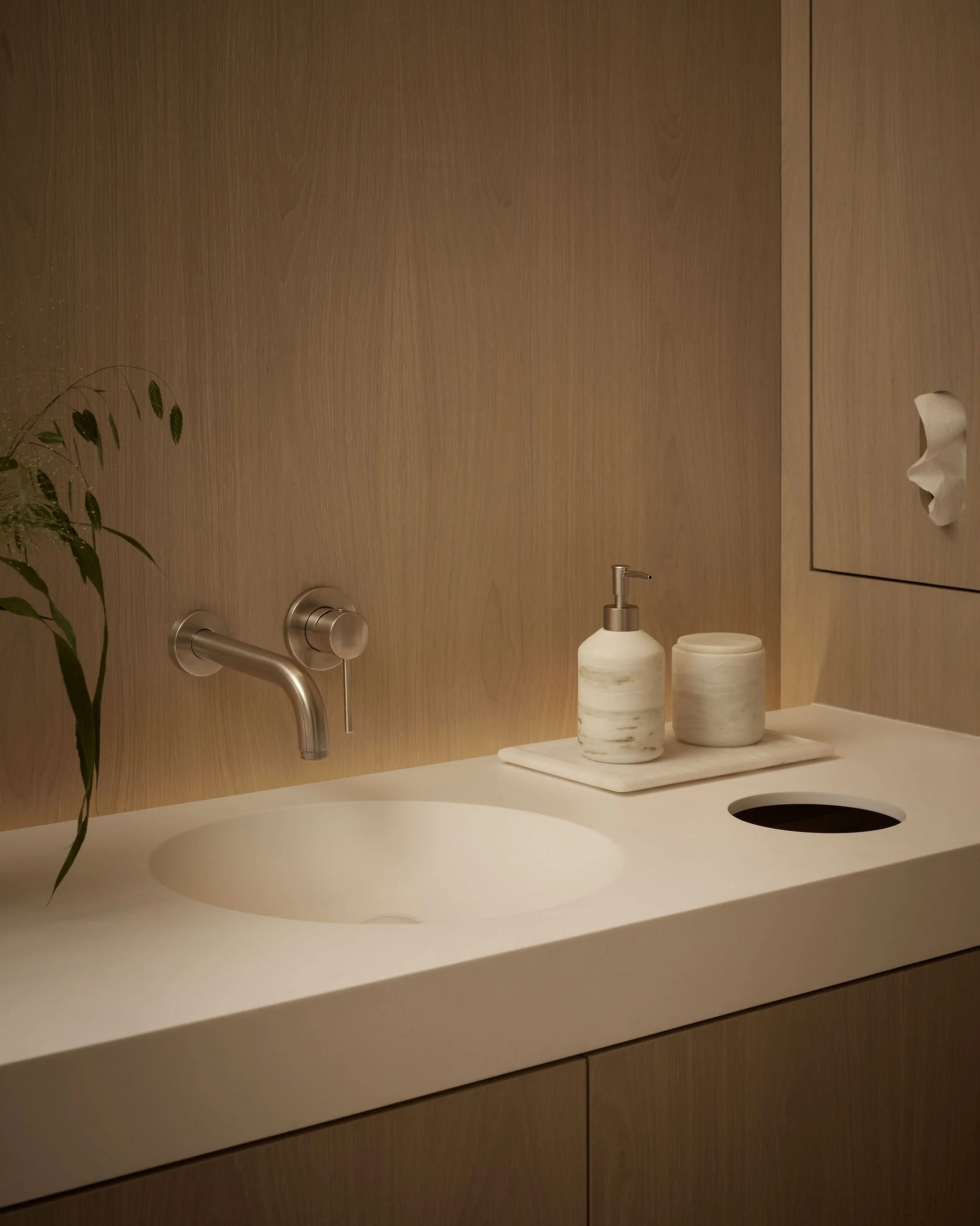 Close up of a minimalist white integrated sink and vanity in a modern wellness clinic, featuring a wall-mounted brushed metal faucet and marble accessories against a sleek wood wall