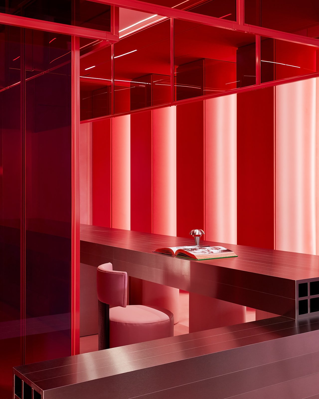 A custom brushed metal cantilevered desk with grid detailing, paired with a plush pink leather stool and a modernist table lamp, set within a bold red and pink anti-clinical wellness space.