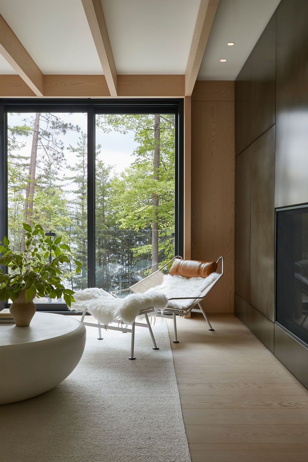 A relaxing corner in a bespoke woodland retreat showcasing floor-to-ceiling windows with panoramic forest views, light wood ceiling beams, and a cozy lounge chair situated next to a custom dark architectural fireplace.