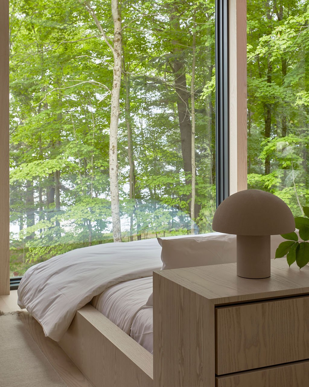 A tranquil bedroom in the Souno recreational residence designed for slow living, featuring a custom wood platform bed, a built-in nightstand with a modern mushroom lamp, and a massive floor-to-ceiling window framing a lush green forest.