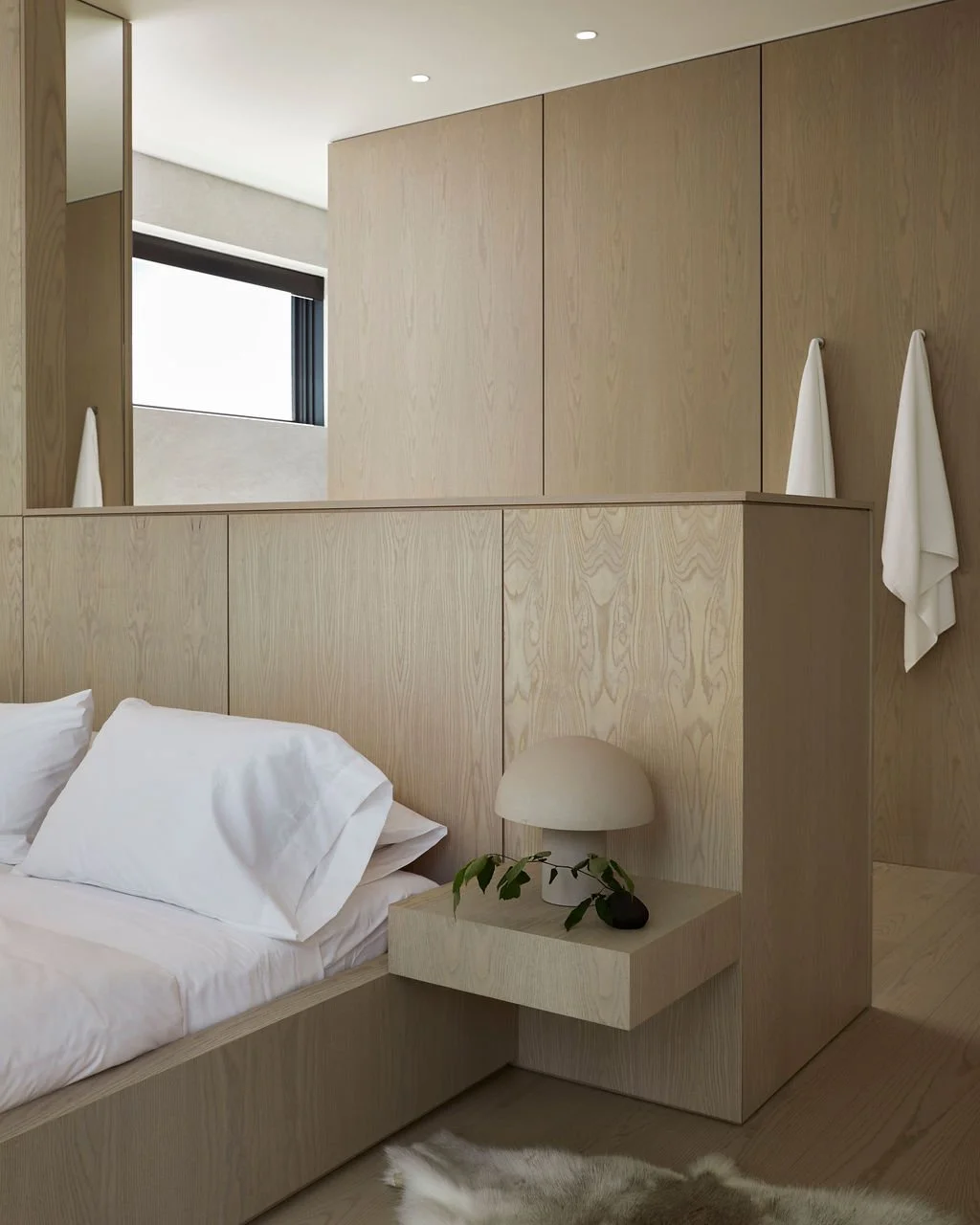 A clever architectural detail in a bespoke cottage showing a custom light wood headboard that functions as a room divider, separating the sleeping area from a dressing space with seamless wooden closets.
