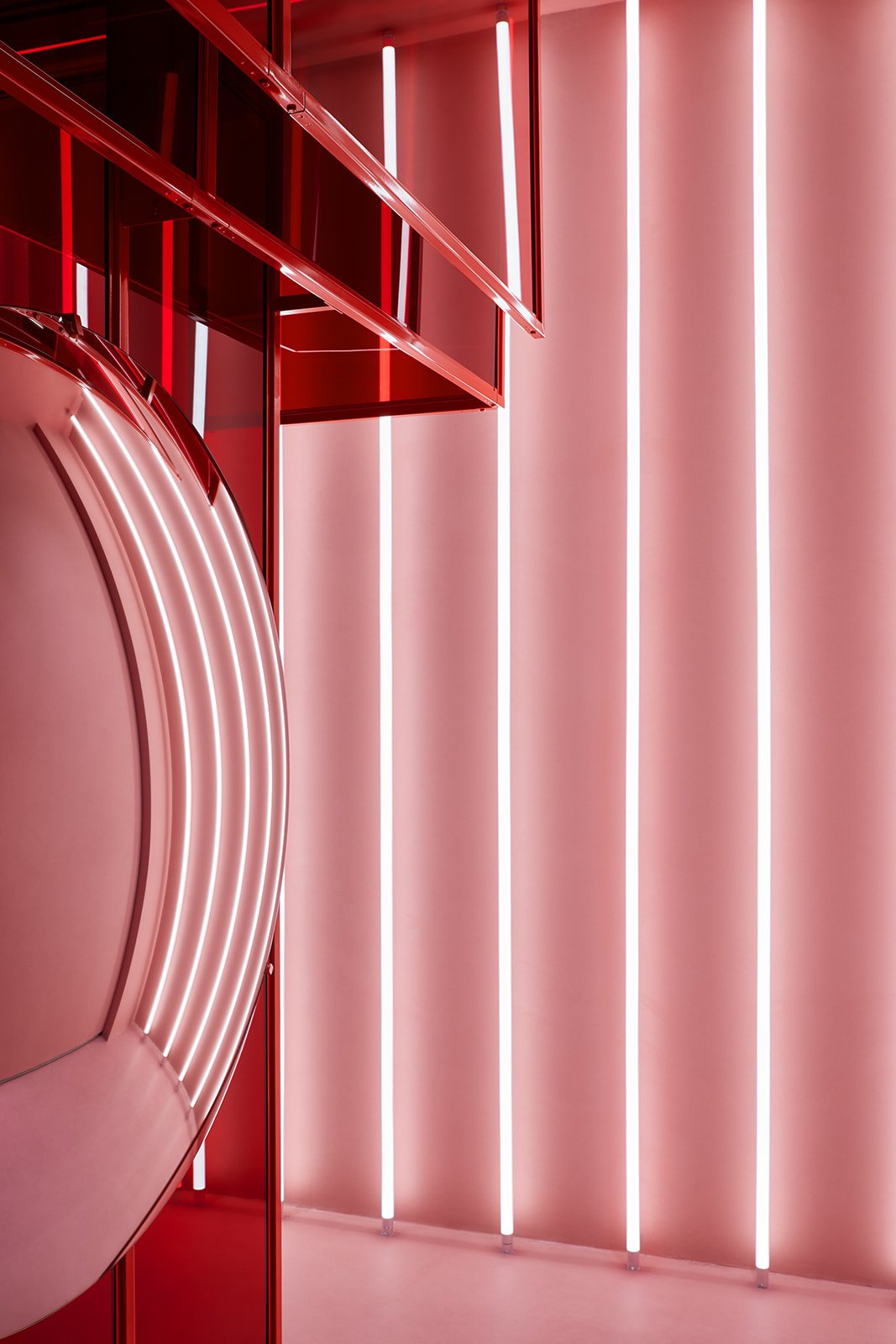 An architectural lighting detail in a luxury medical spa showcasing an illuminated circular feature mirror, red glass framing, and striking vertical LED tube lights against a soft pink wall.
