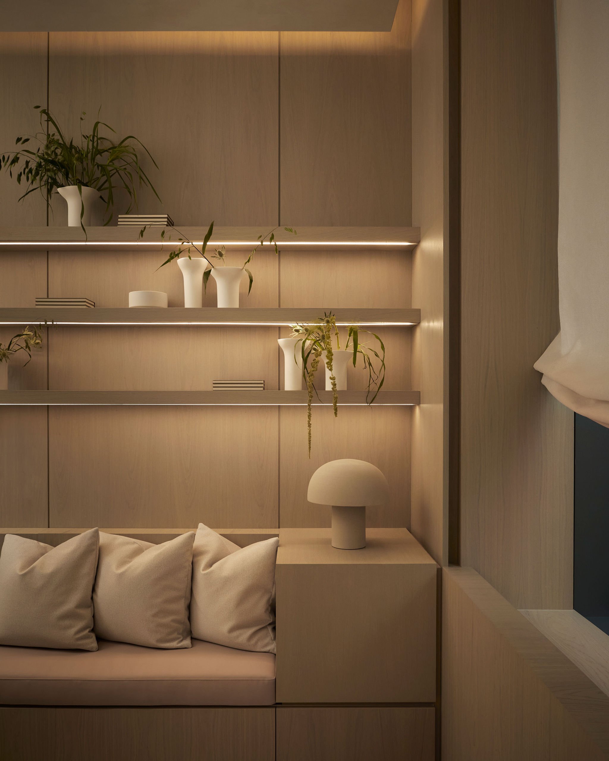 Cozy consultation area in a modern wellness clinic featuring warm wood paneling, a built-in bench with pillows, a sculptural minimalist table lamp, and illuminated floating shelves displaying vases with greenery