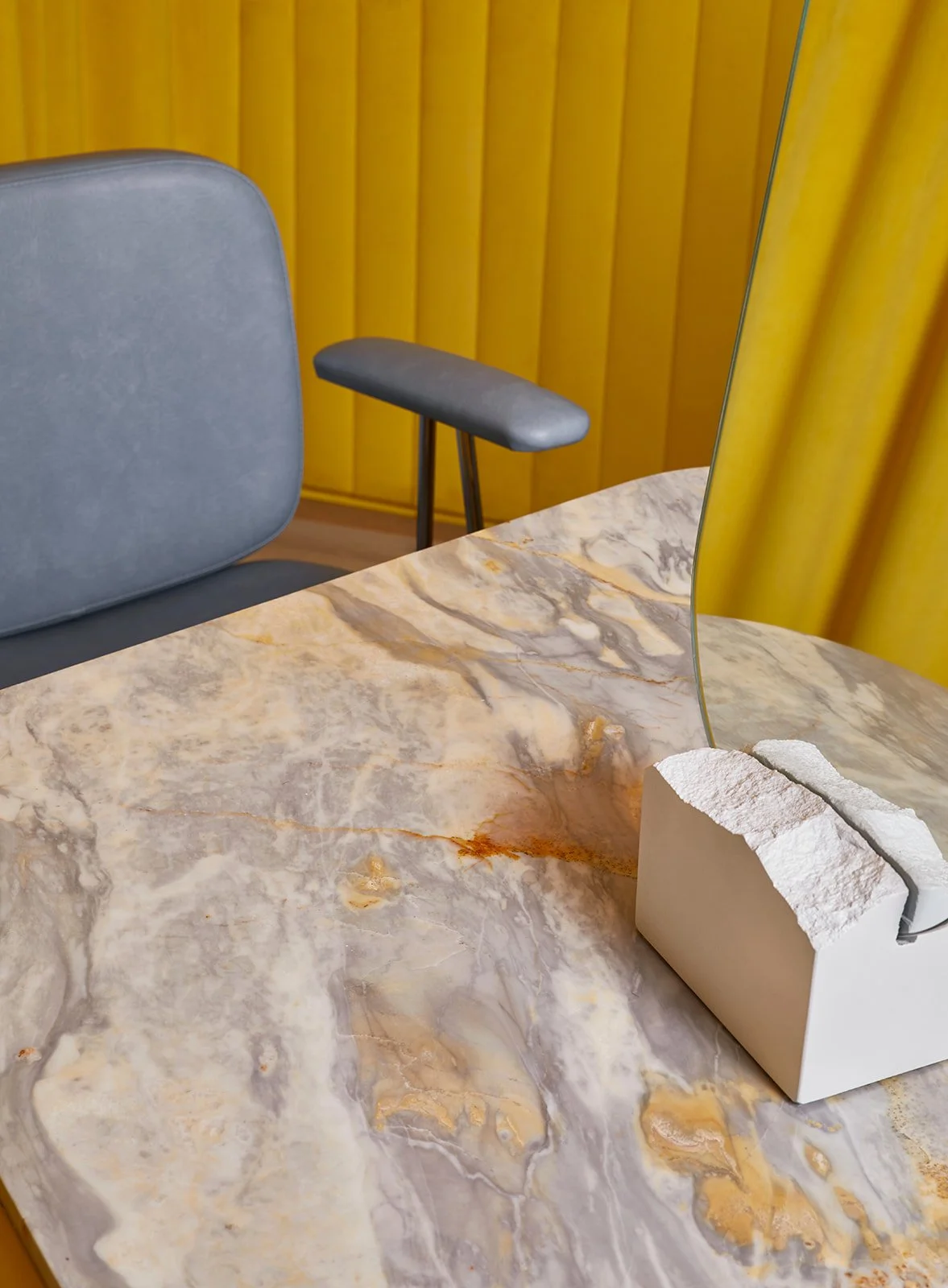 Close-up of a marble table with a modern white tissue holder and a curved glass partition. In the background, a gray chair with a rounded back and a small gray stool with black legs are visible, set against a yellow wooden wall.