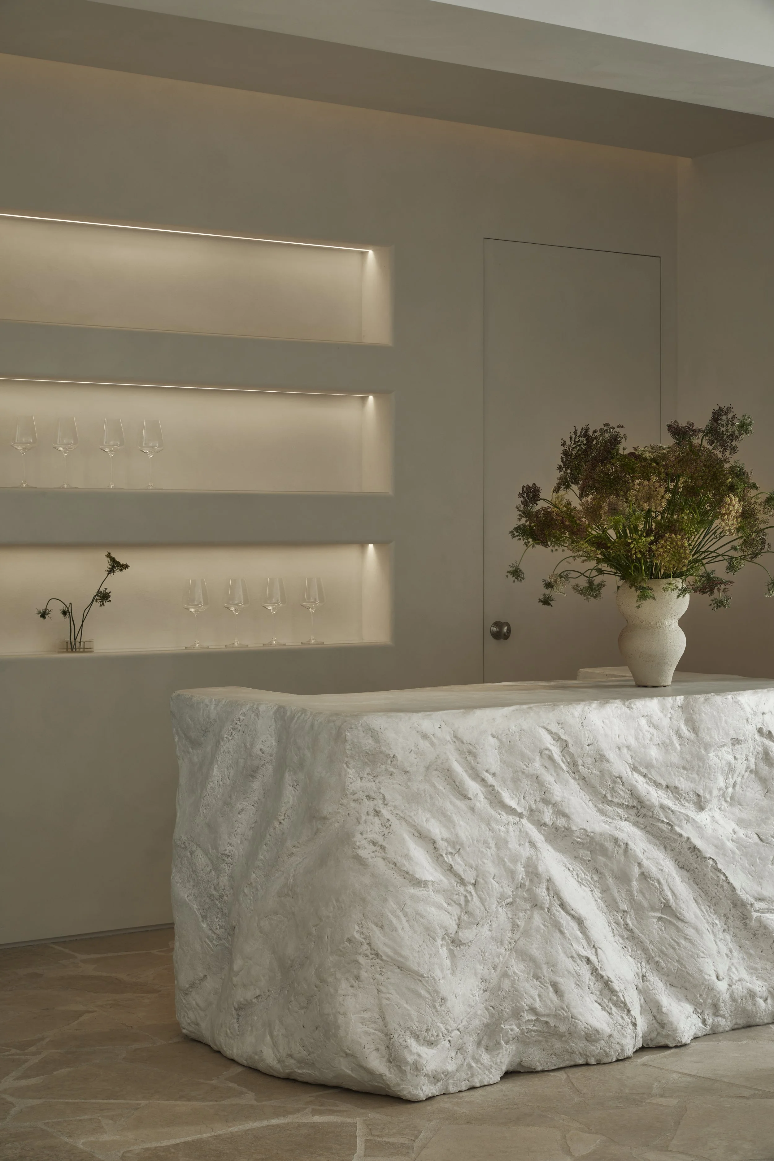 A custom sculpted plaster bar with a raw, rock-like texture in a minimalist hospitality space, featuring built-in illuminated shelving, limewash walls, and a floral arrangement.