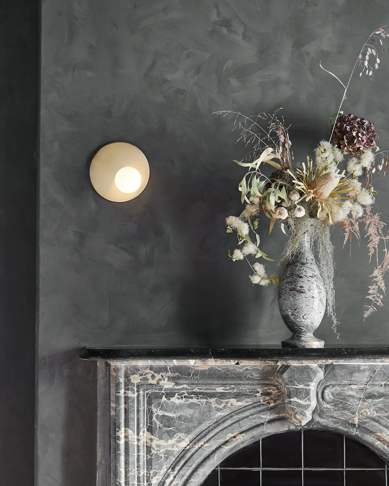 A decorative flower arrangement in a marble vase on a dark mantle against a dark grey textured wall with a modern wall sconce