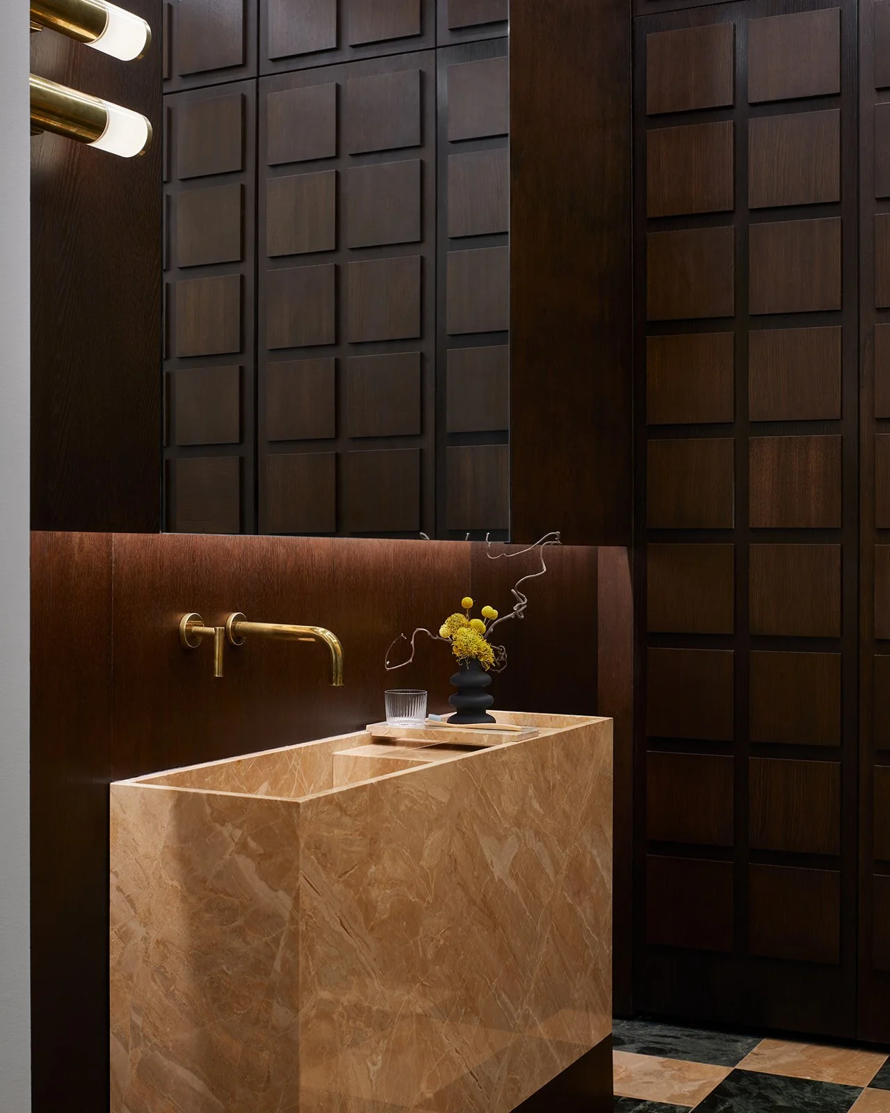 A luxurious clinical hand-washing station featuring a custom monolithic tan marble sink, wall-mounted brass faucets, and rich dark wood grid-paneled walls that completely conceal the clinical storage.
