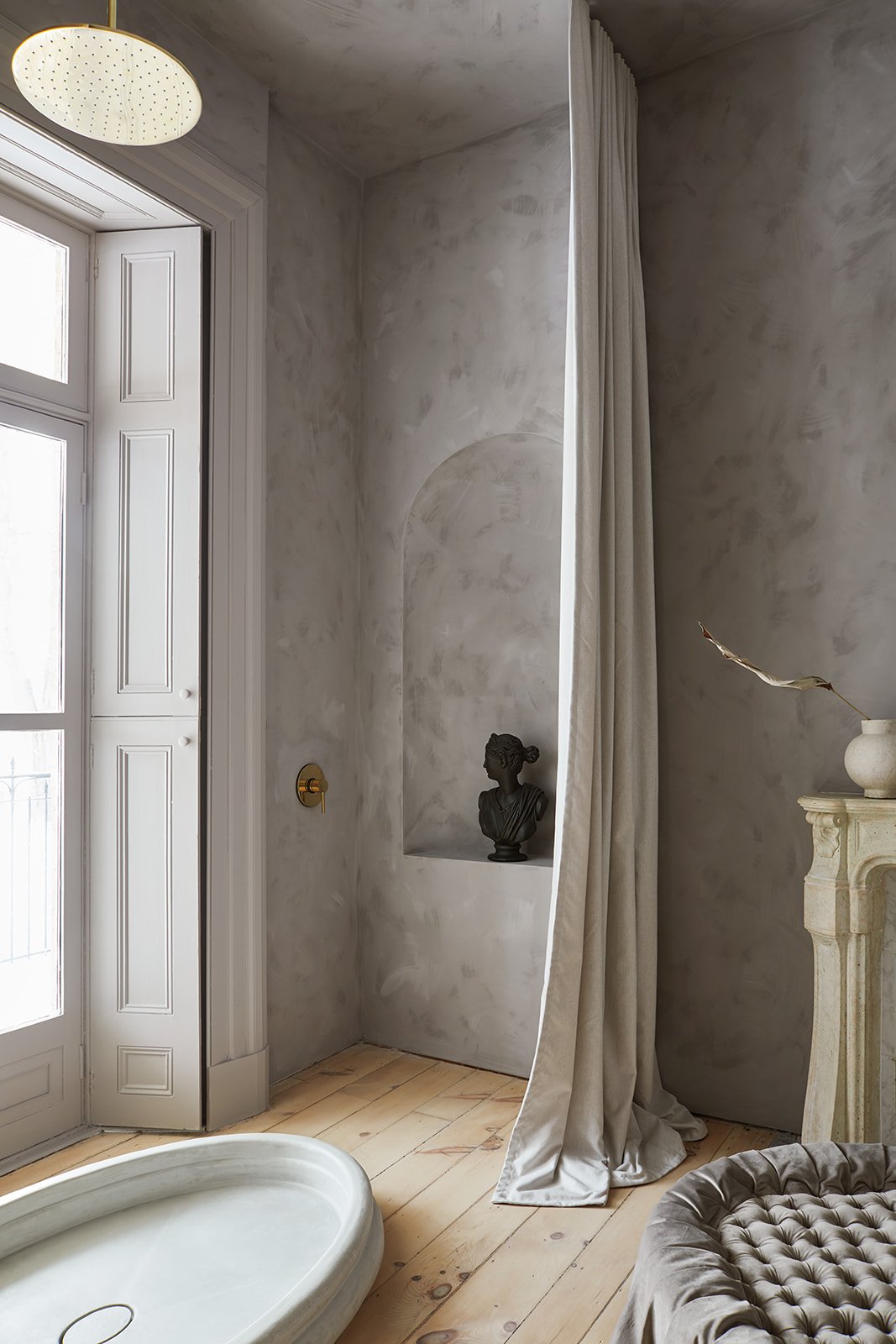 A bathroom with a round rain shower head, a large window, a small bust sculpture on a nook, a curtain, and a stone fireplace with a vase and dried leaf.