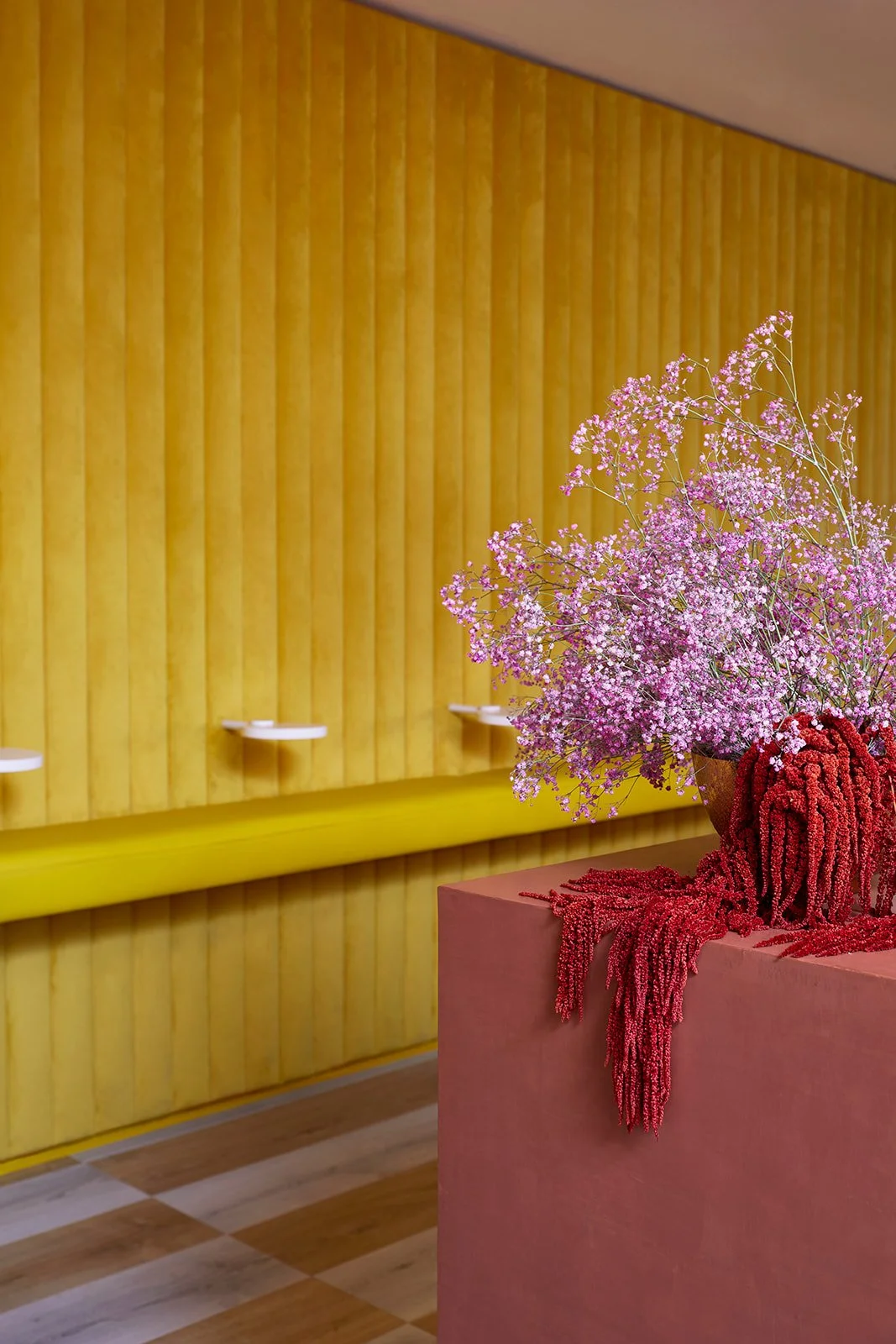 Pink flowers in a red vase on a pink pedestal against a yellow wall with white shelves.