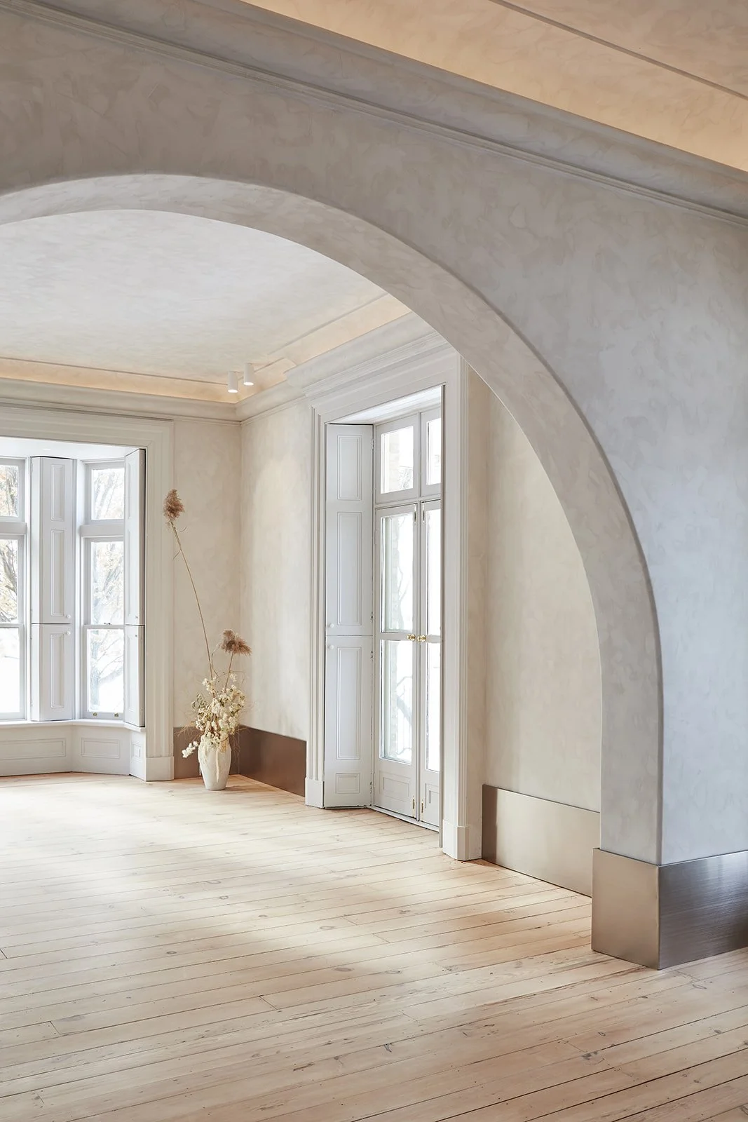 Bright, empty room with large bay windows, light-colored wooden floor, cream textured walls, decorative arch, and a tall vase with dried flowers