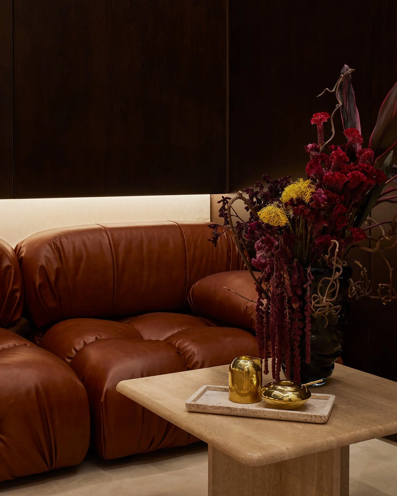 A warm, restorative consultation and waiting lounge in a boutique dental office, showcasing a plush, tufted caramel leather sofa, a custom travertine stone coffee table, and rich dark wood walls.
