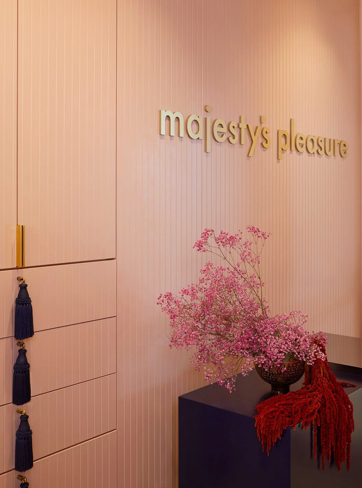 Pink wall with gold lettering that says 'majesty's pleasure'. A dark table with a vase of pink flowers and a red decorative cloth, and storage drawers with dark blue tassels.