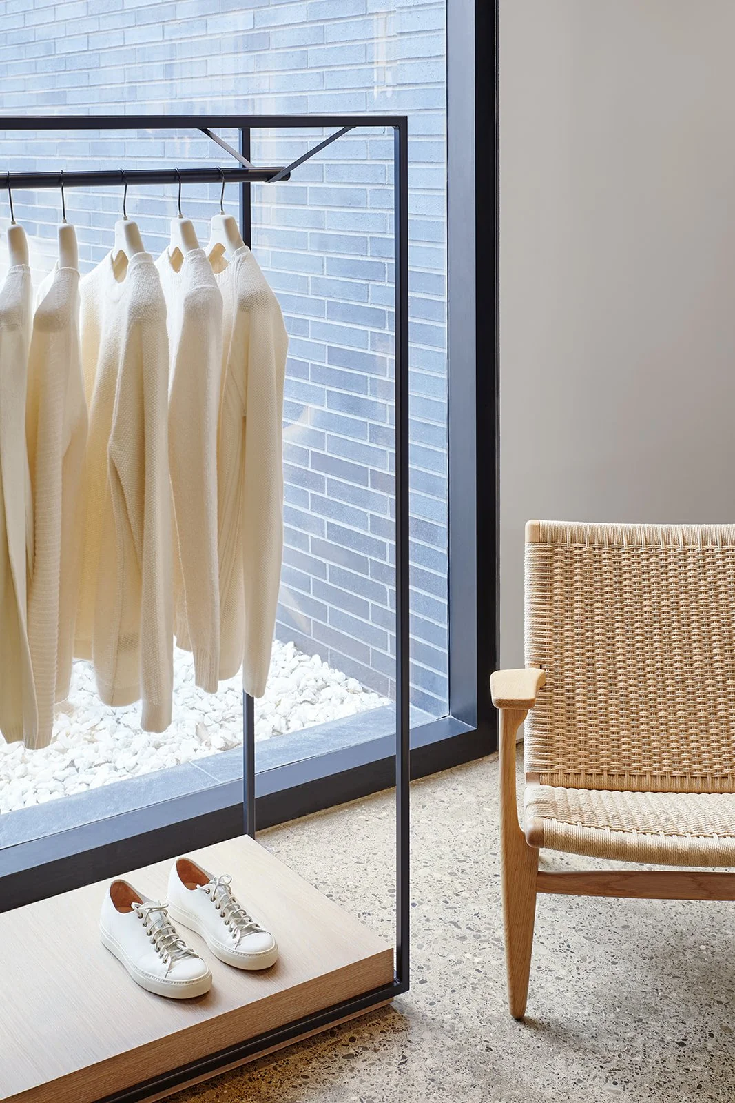 Cream-colored sweaters hanging on a black metal clothing rack near a window, with a pair of white sneakers on a wooden platform nearby, and an armchair with woven upholstery beside them in a modern room.