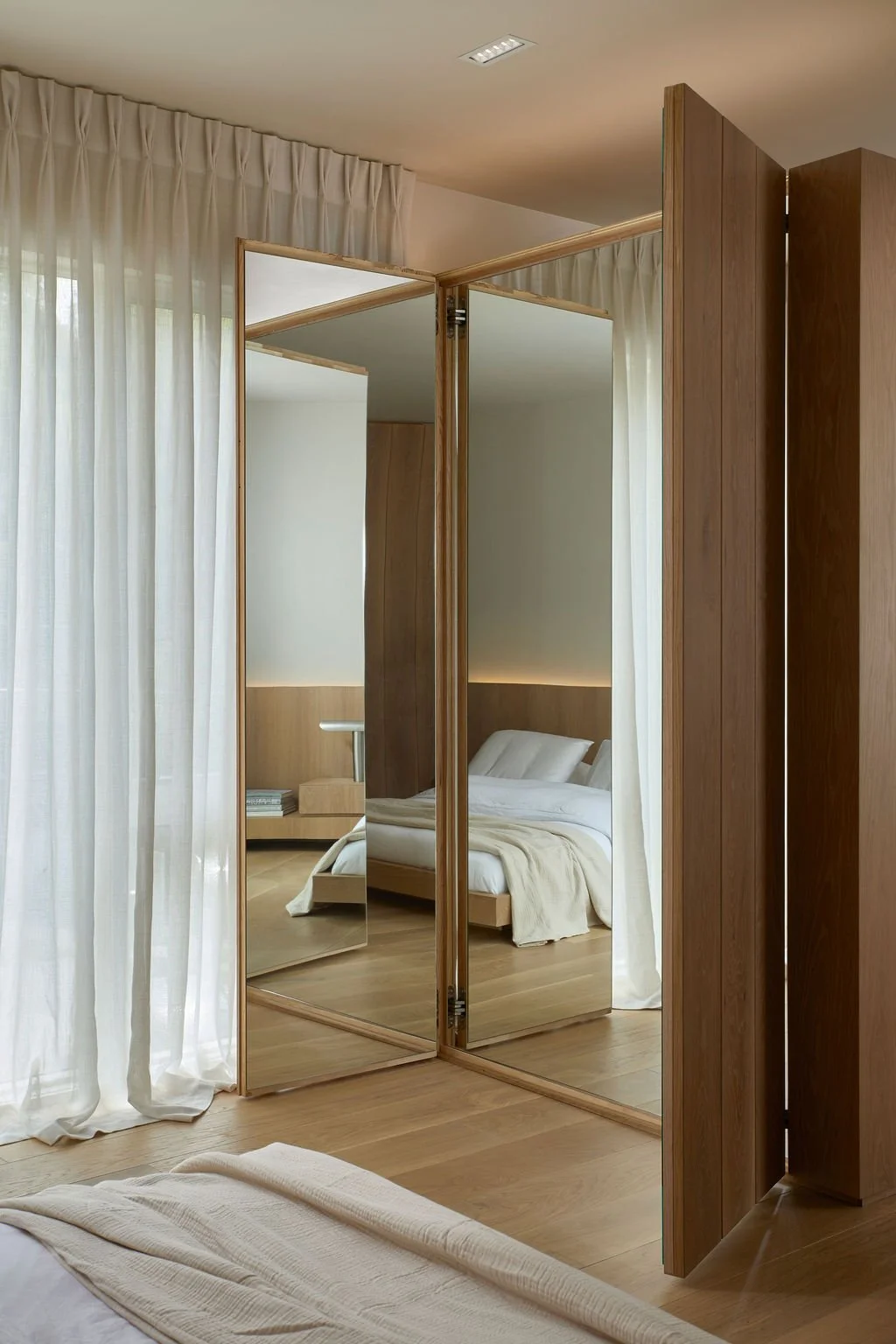 Custom bi-fold mirrored closet doors set seamlessly within ribbed oak wall paneling, standing partially open to reflect a serene, minimalist luxury bedroom and sheer window drapery.