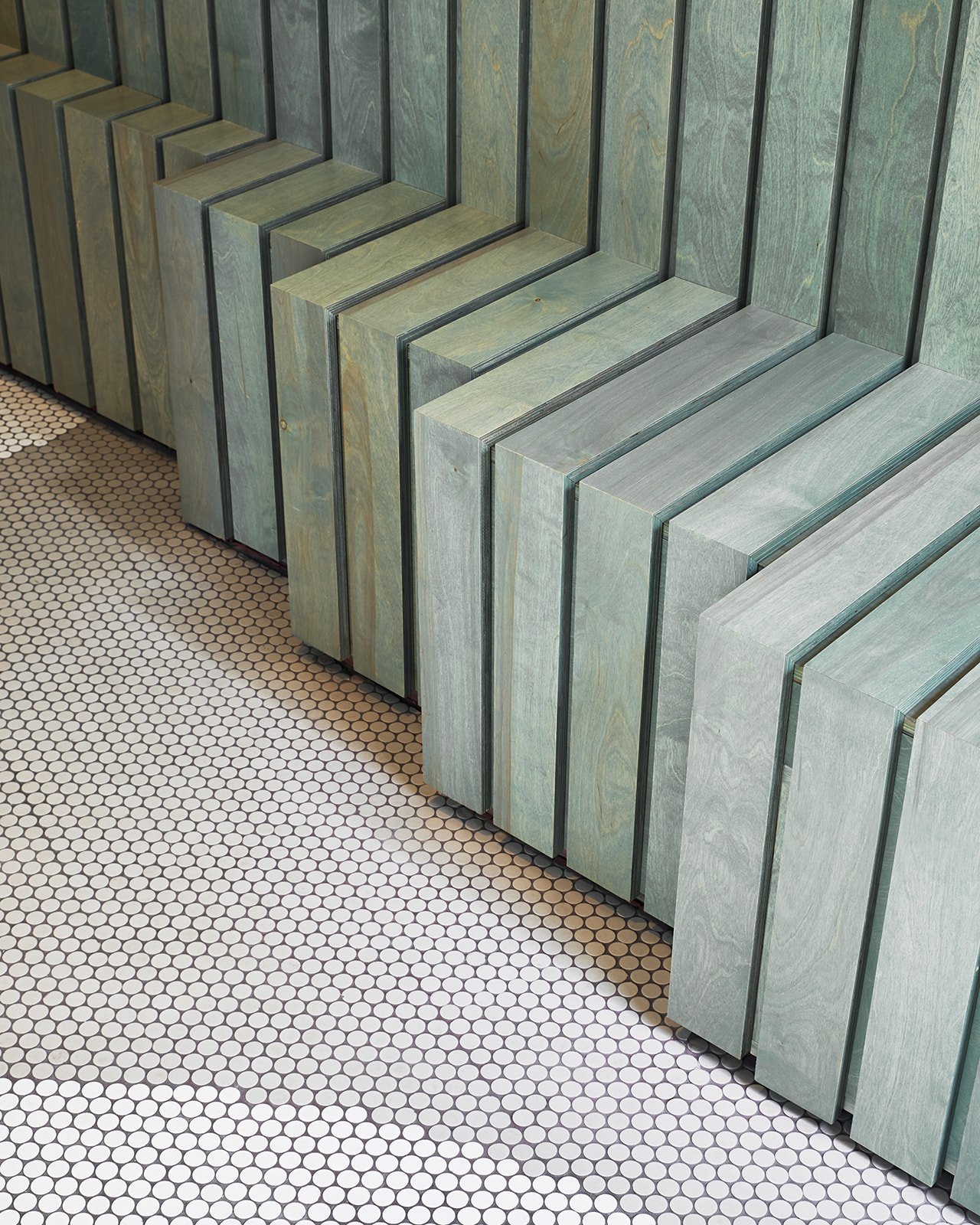 A row of vertically standing greenish wood planks against a wall with small round white tile flooring.