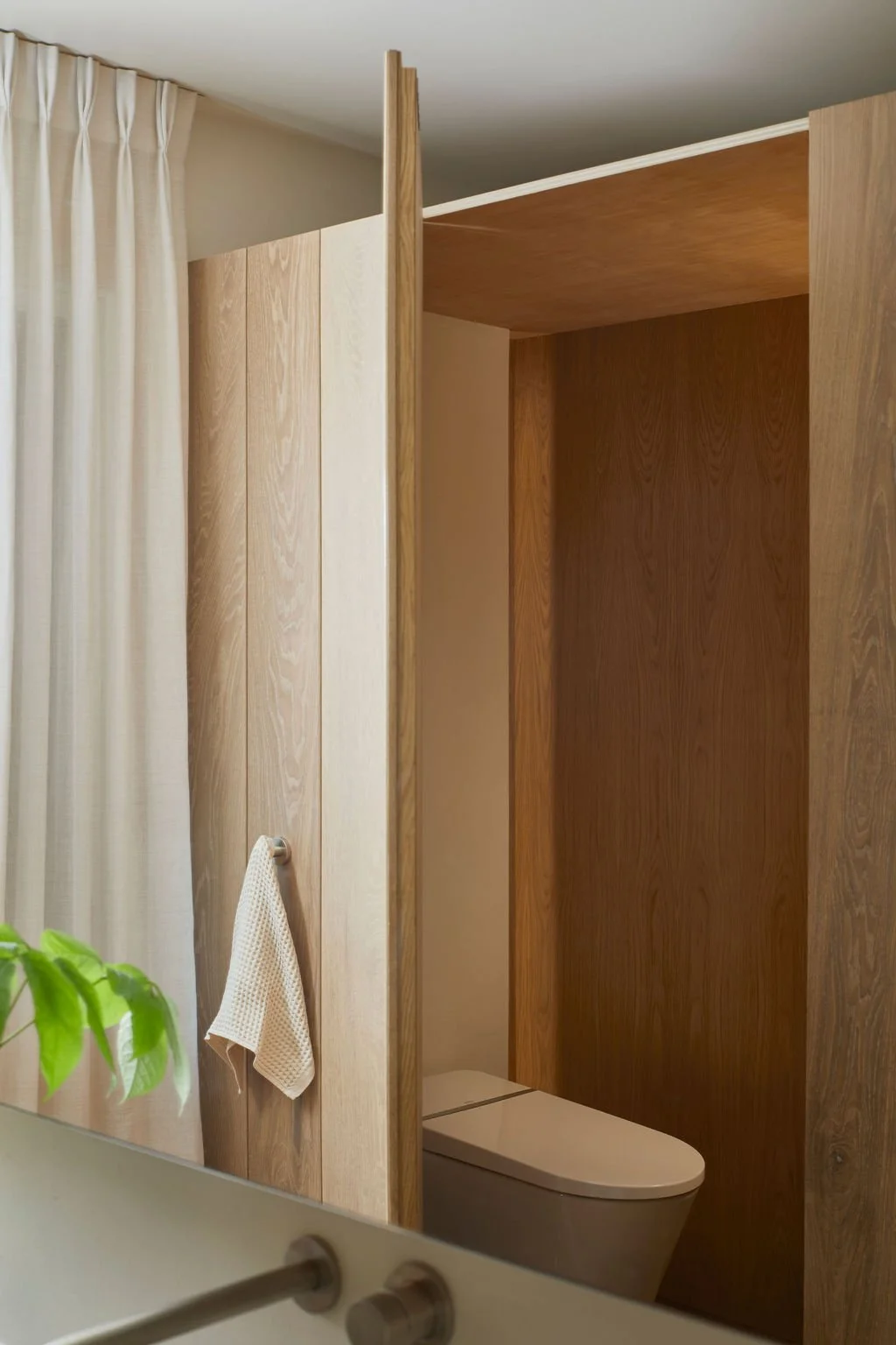 A minimalist water closet discreetly concealed behind a custom ribbed oak architectural door, situated next to a window with sheer white drapery in a tranquil Toronto home.