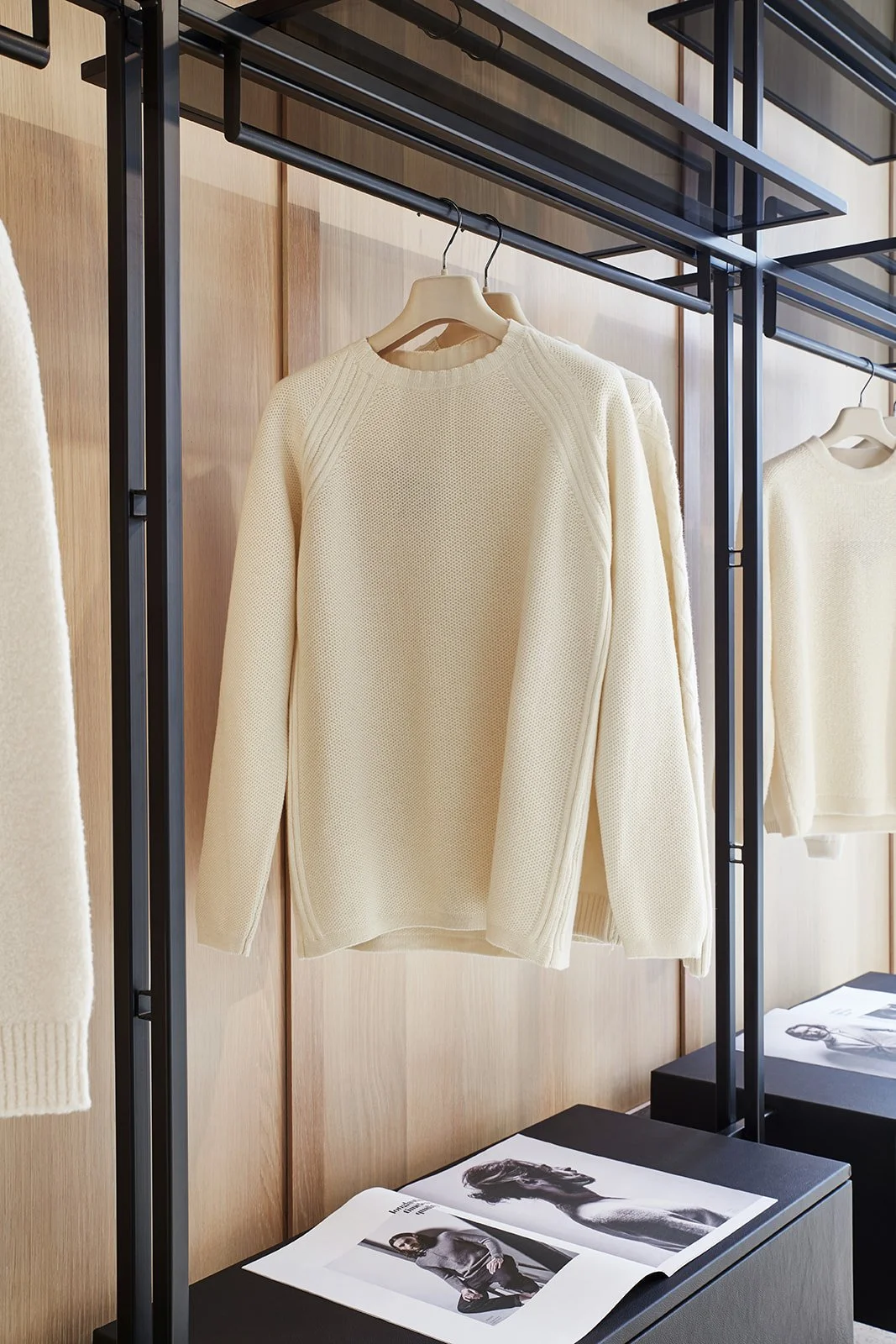 Cream-colored knit sweater hanging on a black metal clothing rack in a retail store with wooden wall panels and fashion magazines on a black surface below.