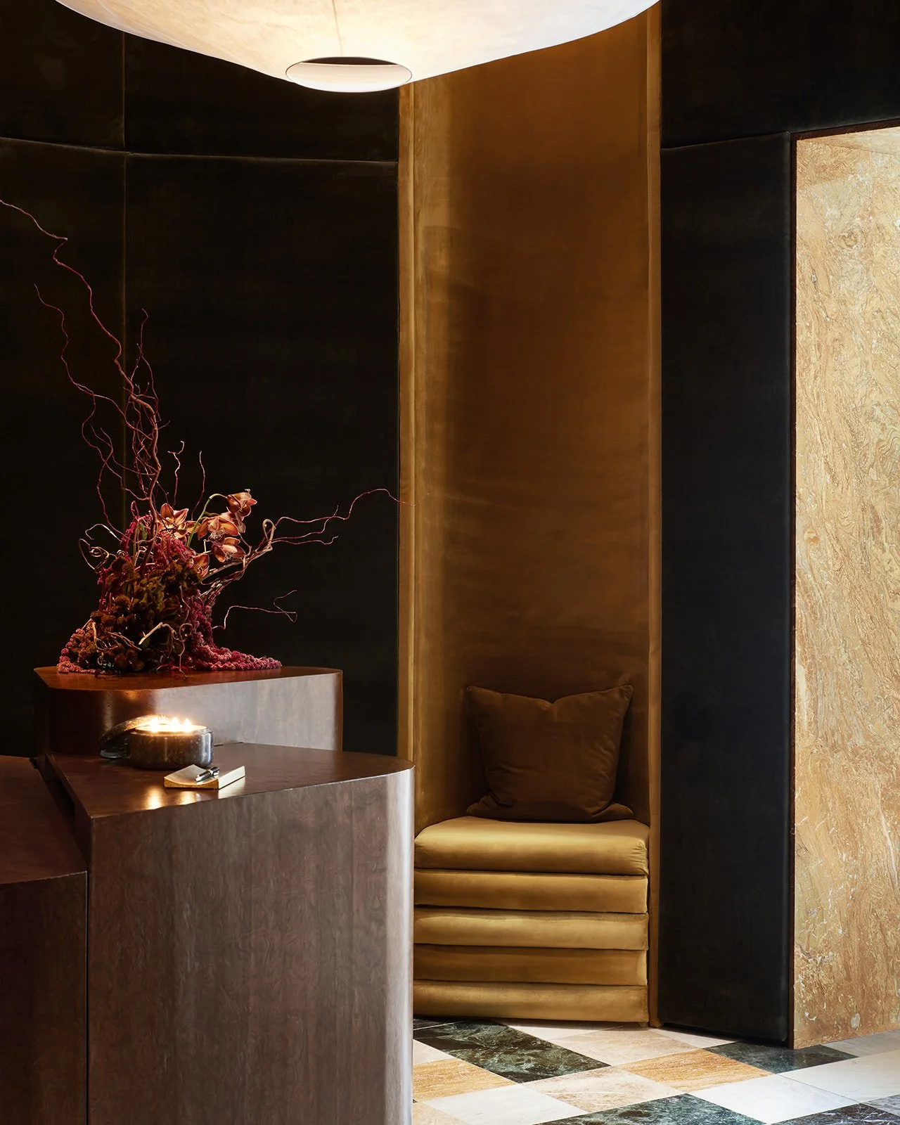 A cozy, anti-clinical waiting nook built into dark wood paneled walls, featuring plush gold velvet seating next to a bespoke wood reception desk and striking marble flooring.