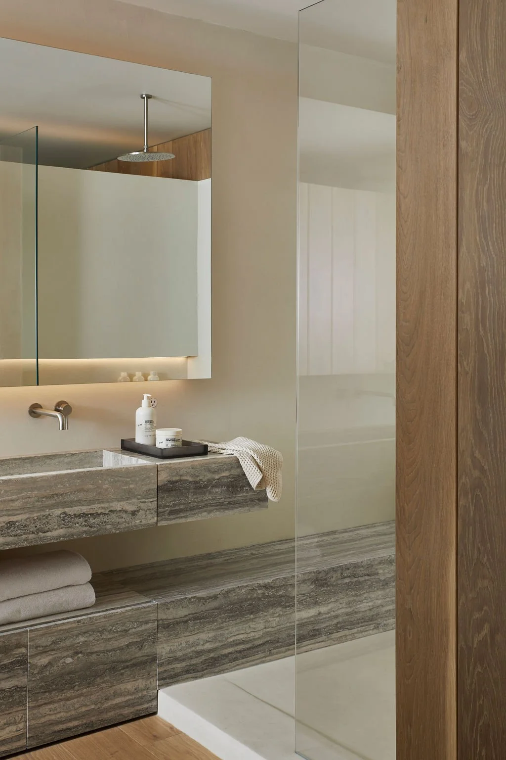 A close-up of a bespoke floating travertine stone bathroom sink and lower storage shelf, seamlessly intersecting with a minimalist frameless glass shower enclosure in a luxury residential suite.