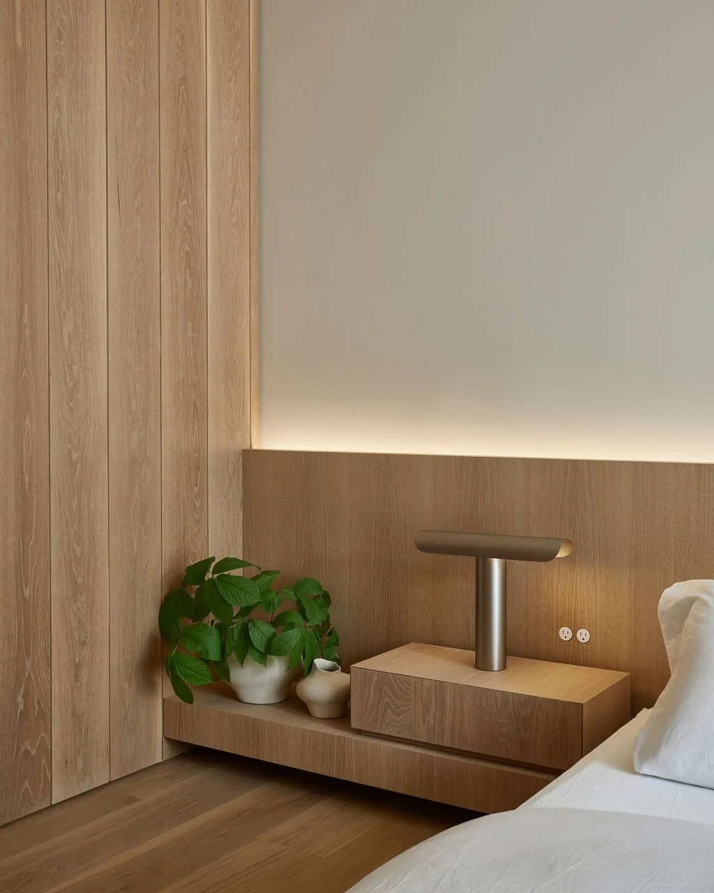 An architectural detail of a custom floating oak nightstand integrated into a ribbed wood headboard, styled with a modern metal table lamp and a potted plant bathed in warm LED lighting.