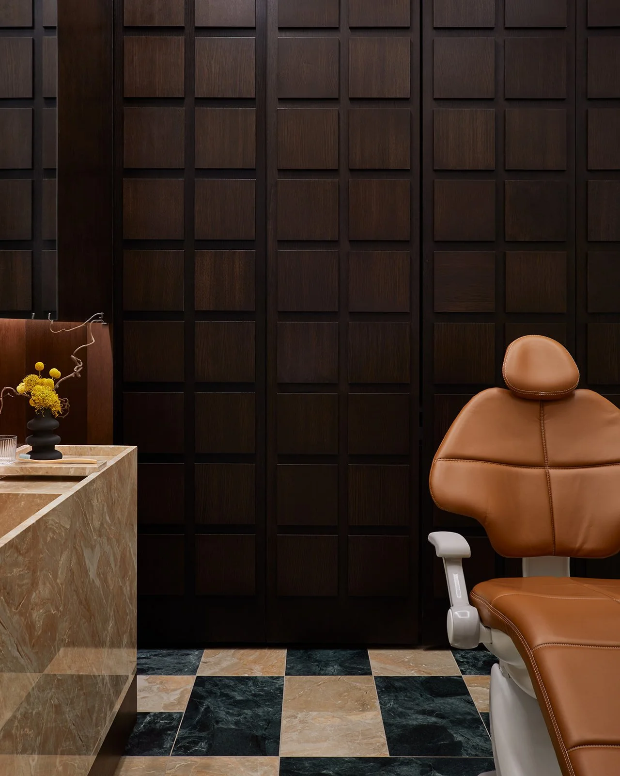 An elevated dental treatment room featuring a premium caramel leather dental chair, striking marble checkerboard flooring, and floor-to-ceiling dark wood grid millwork designed to reduce patient anxiety.