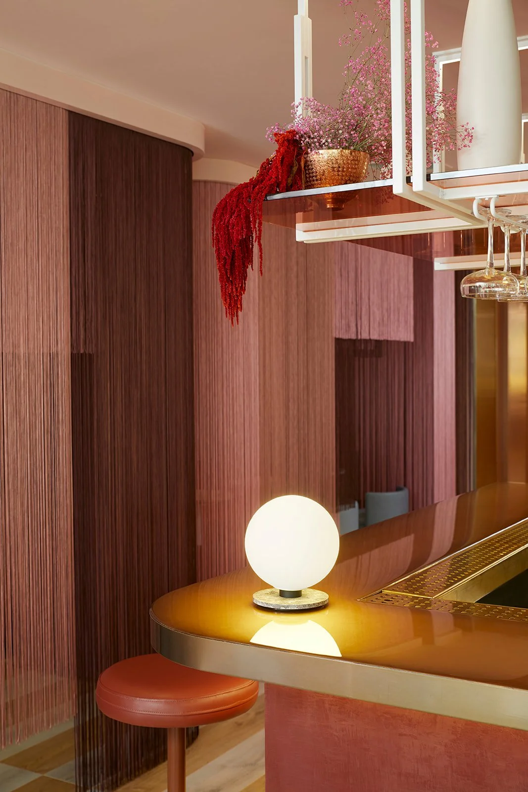 A modern bar area with wooden wall panels, a curved wooden countertop with a spherical white lamp on it, and a red bar stool. Decor includes pink and red flowers in vases and hanging wine glasses.