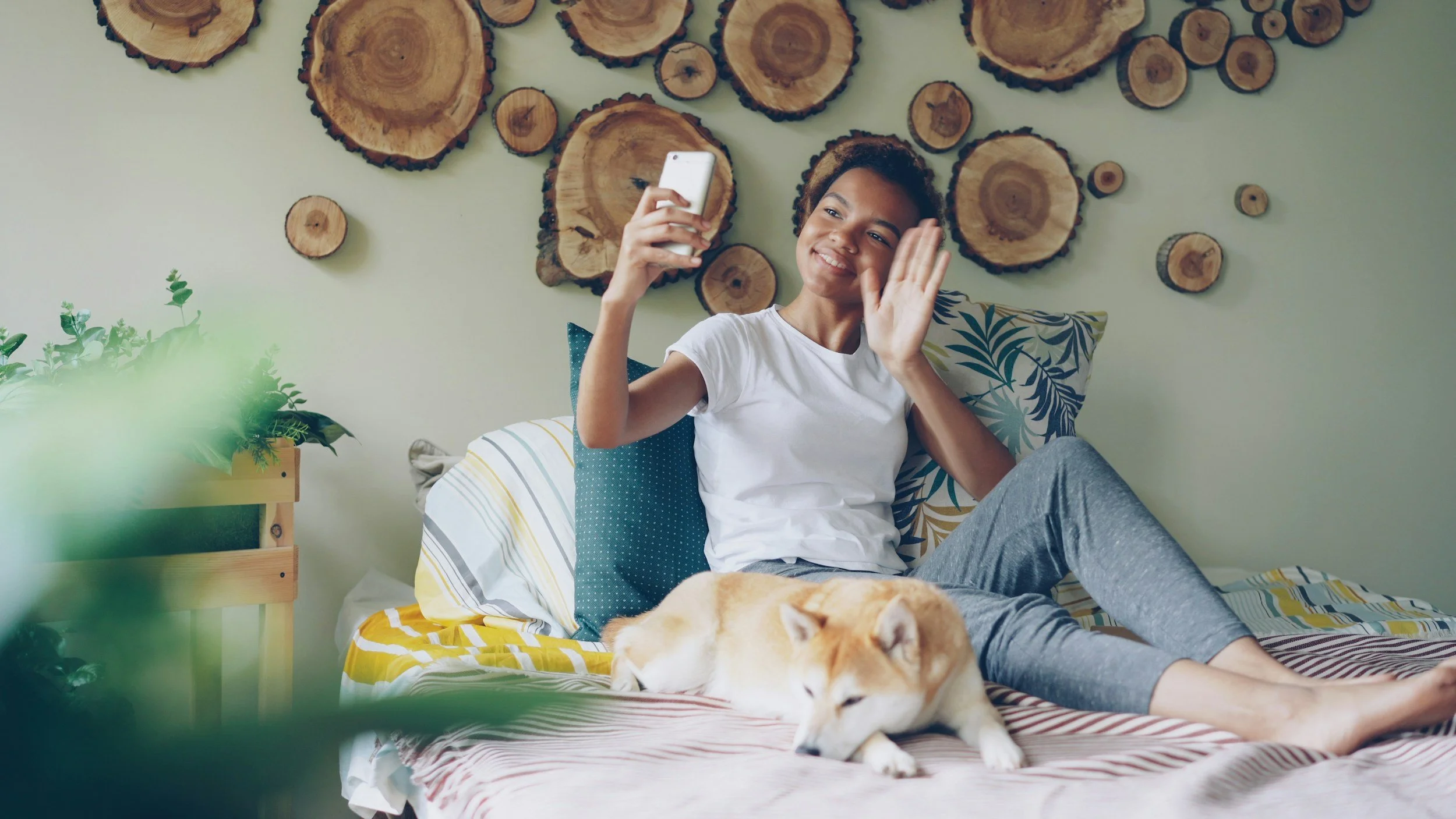 Young woman sitting on a bed waves hello into a phone camera as a dog lies next to her
