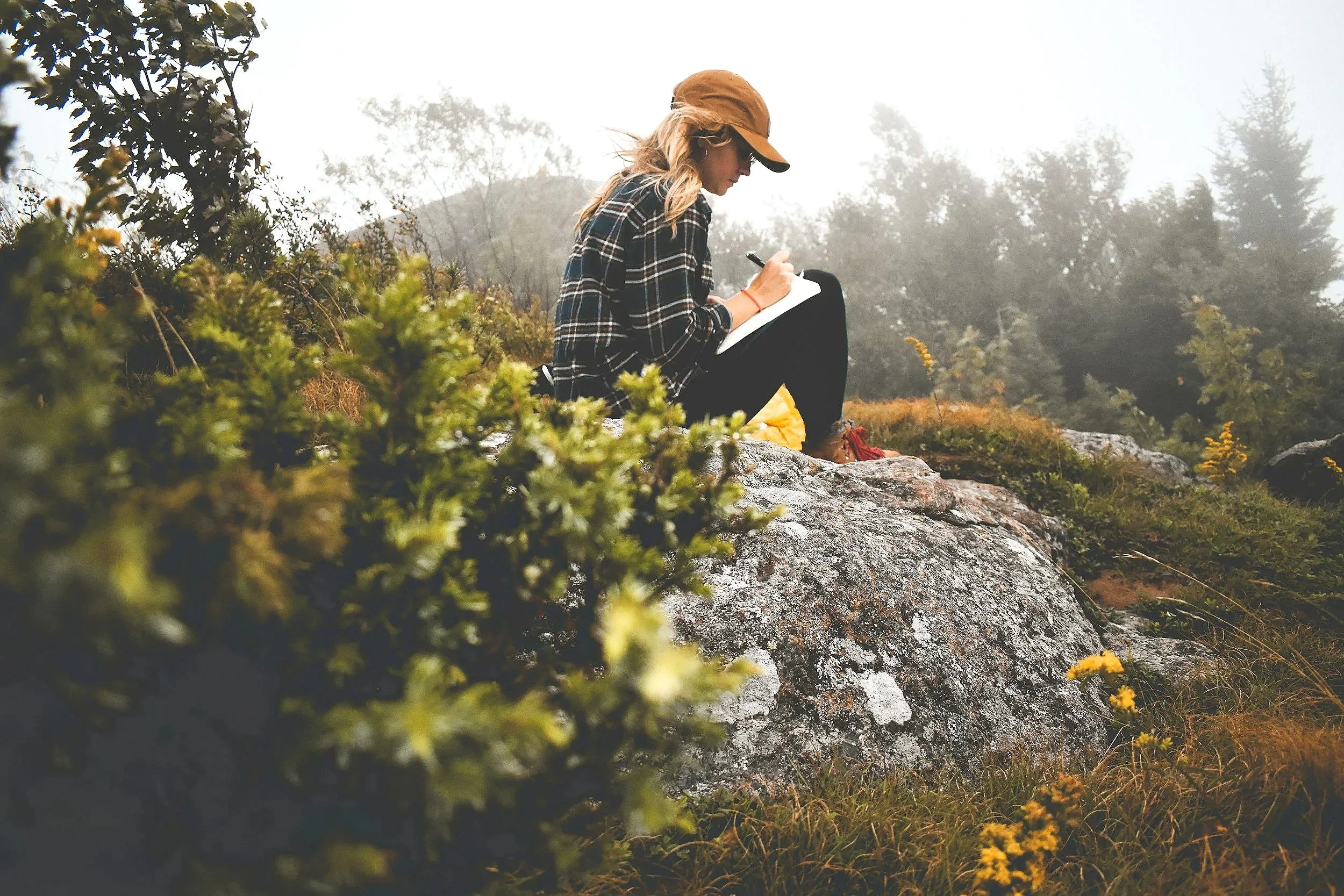 Woman sits on rock in nature and writes in her journal