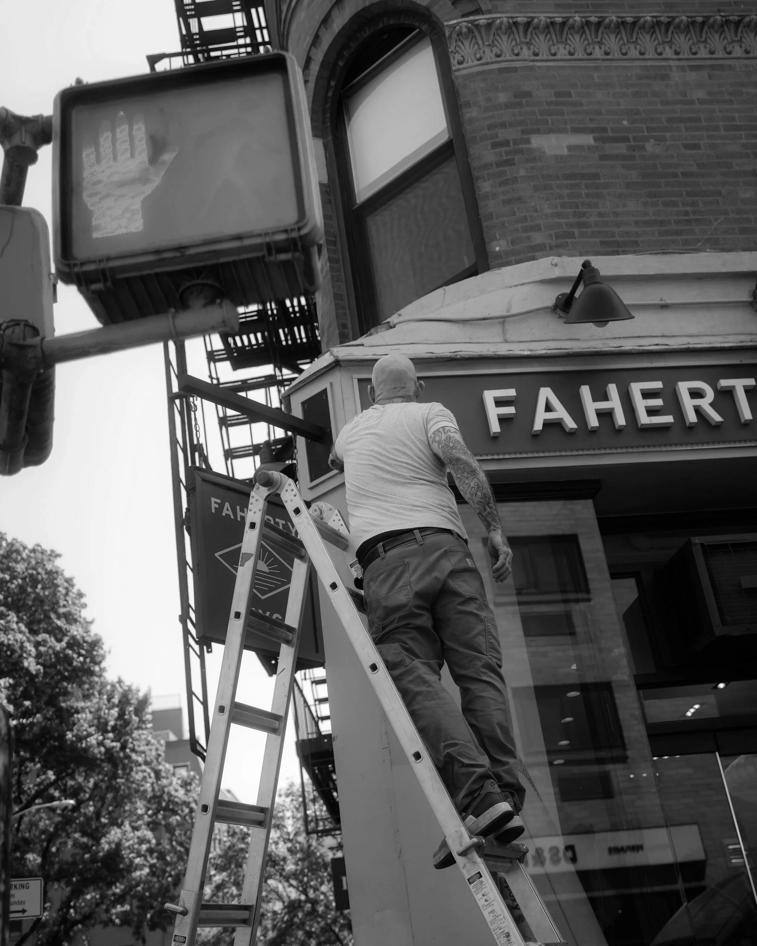A man standing on a ladder working on the exterior of a building. There is a sign with the name 'FAHERTY' on the building. A stage light is mounted on the building's exterior. Part of a fire escape ladder and sidewalk signs are also visible.