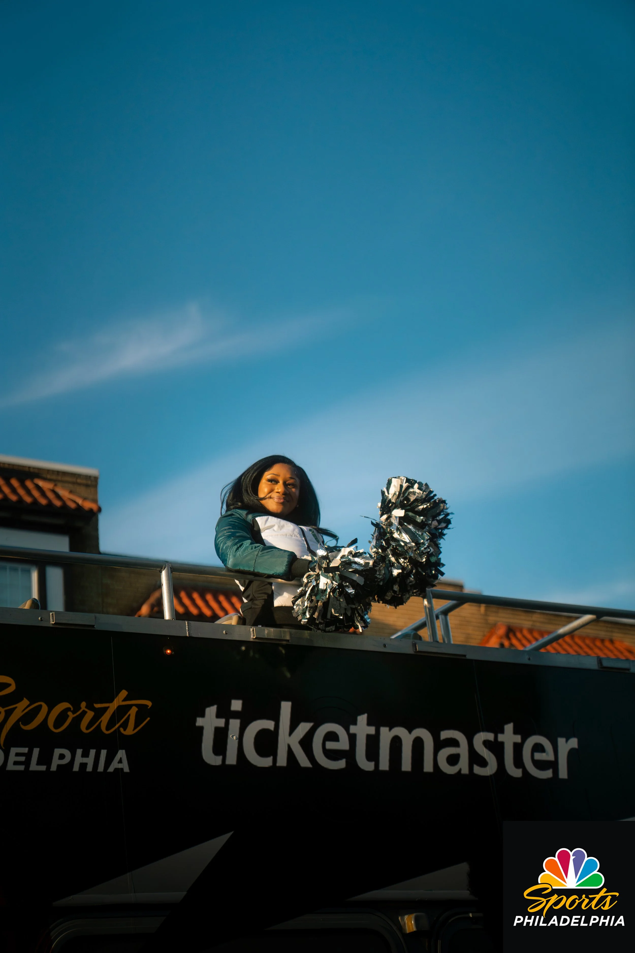 A female cheerleader standing on a float at a parade, holding pom-poms, with a clear blue sky overhead and buildings in the background.