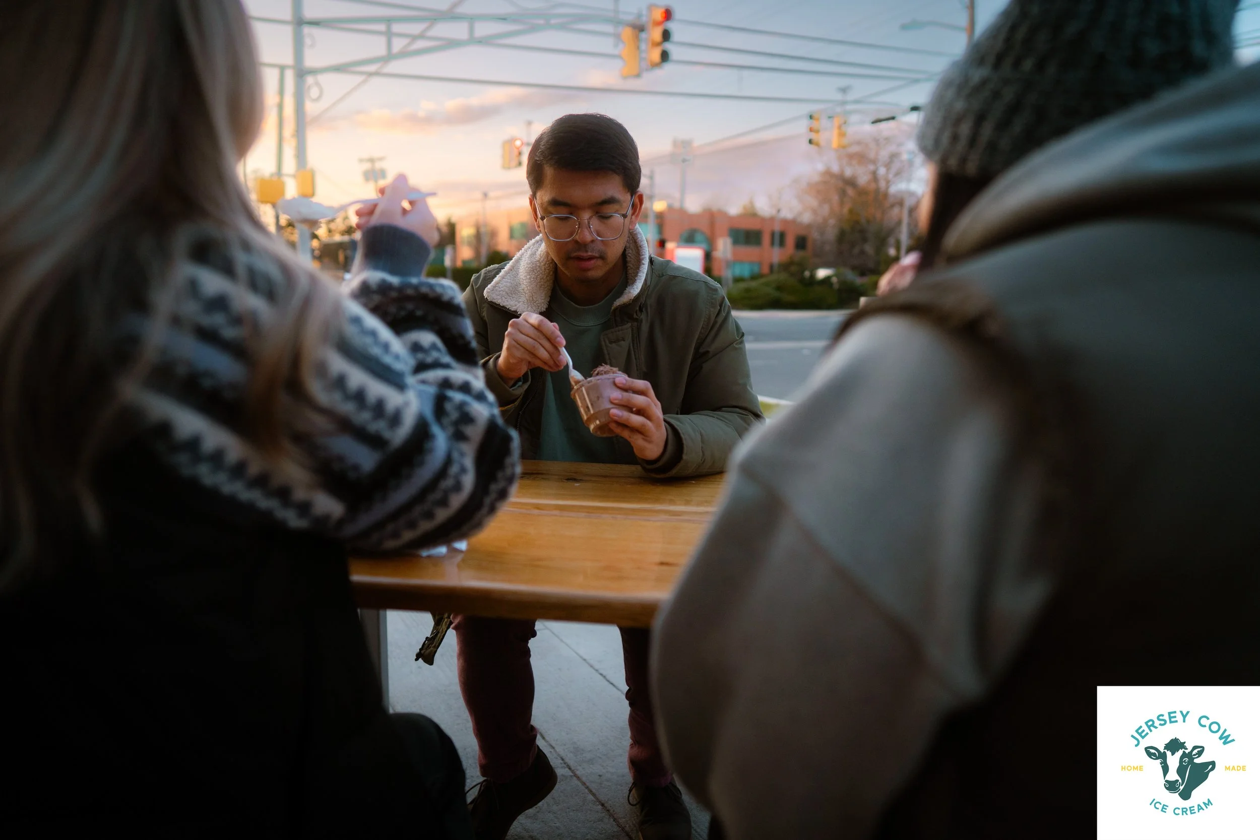 A young man with glasses and a green jacket is eating ice cream while sitting at a table outside. Two women are partially visible on either side of him, one with long hair and the other wearing a knit hat. The background shows traffic lights and a su