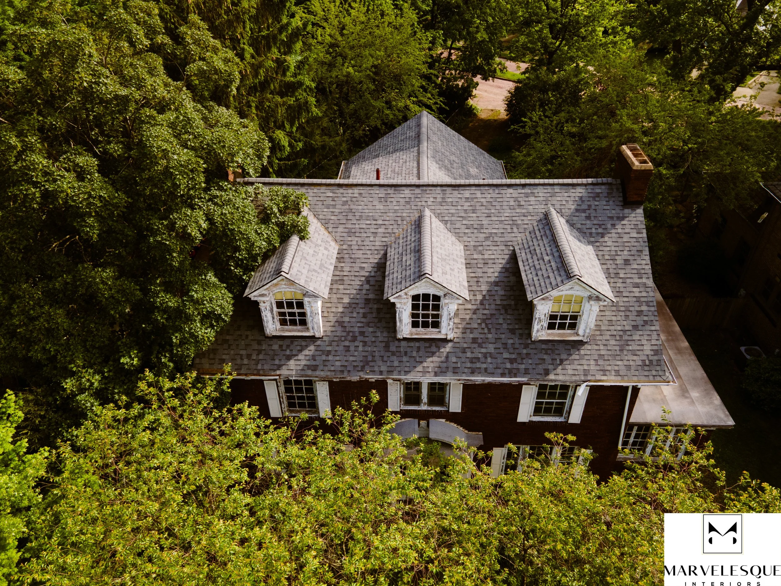 An aerial view of a large house with a gray shingled roof, surrounded by tall green trees and a driveway visible in the background.