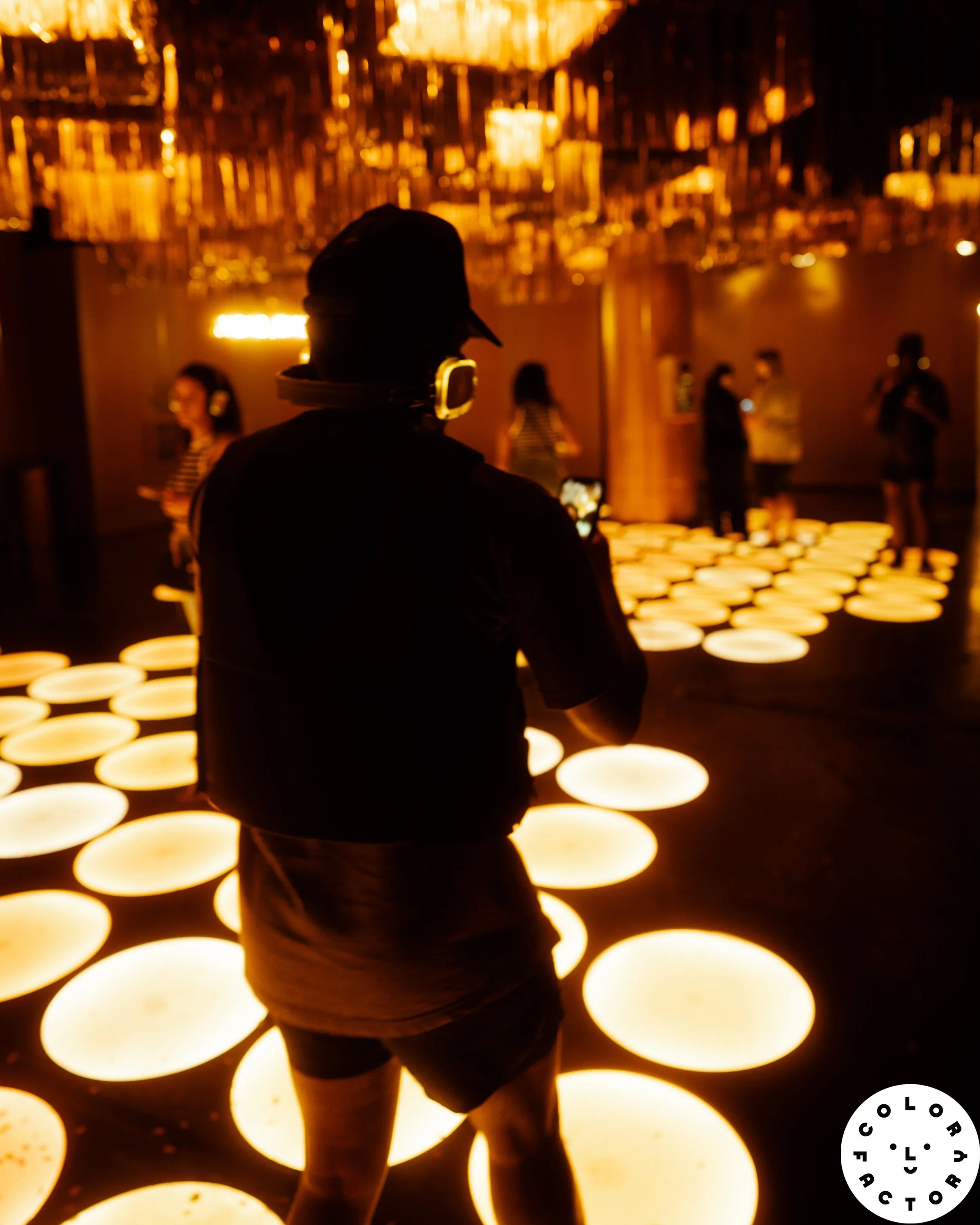 Person with headphones and a baseball cap standing in front of a glowing wall of circular lights in a dark room.