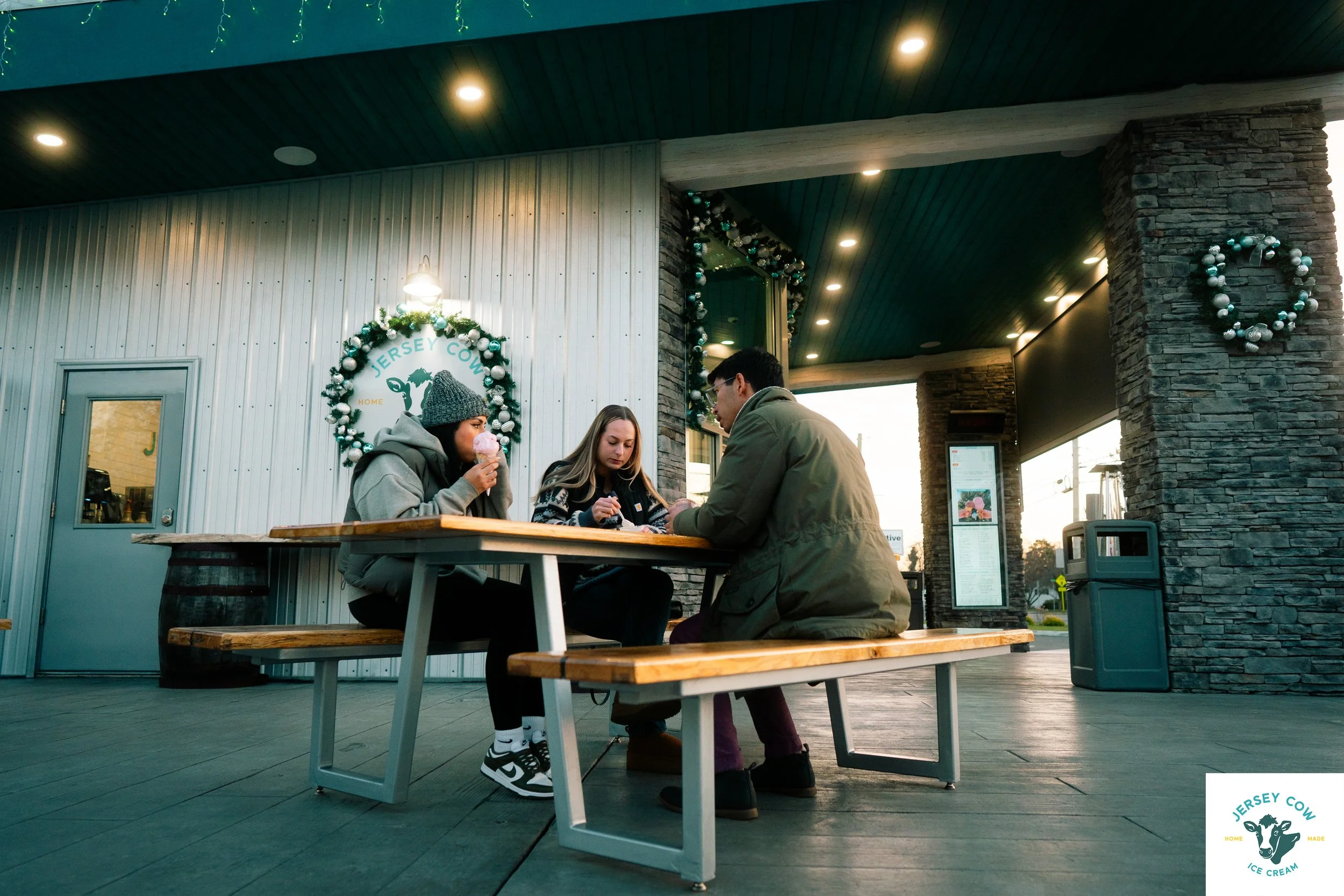 Three people sitting at a wooden picnic table outside a building decorated with Christmas wreaths and lights, with one person eating ice cream cone and others looking at their phones.