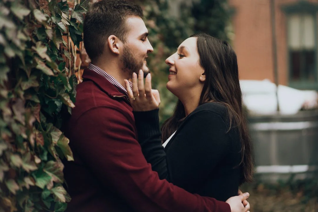 A couple faces each other closely, smiling, with a green leafy wall behind them outdoors.