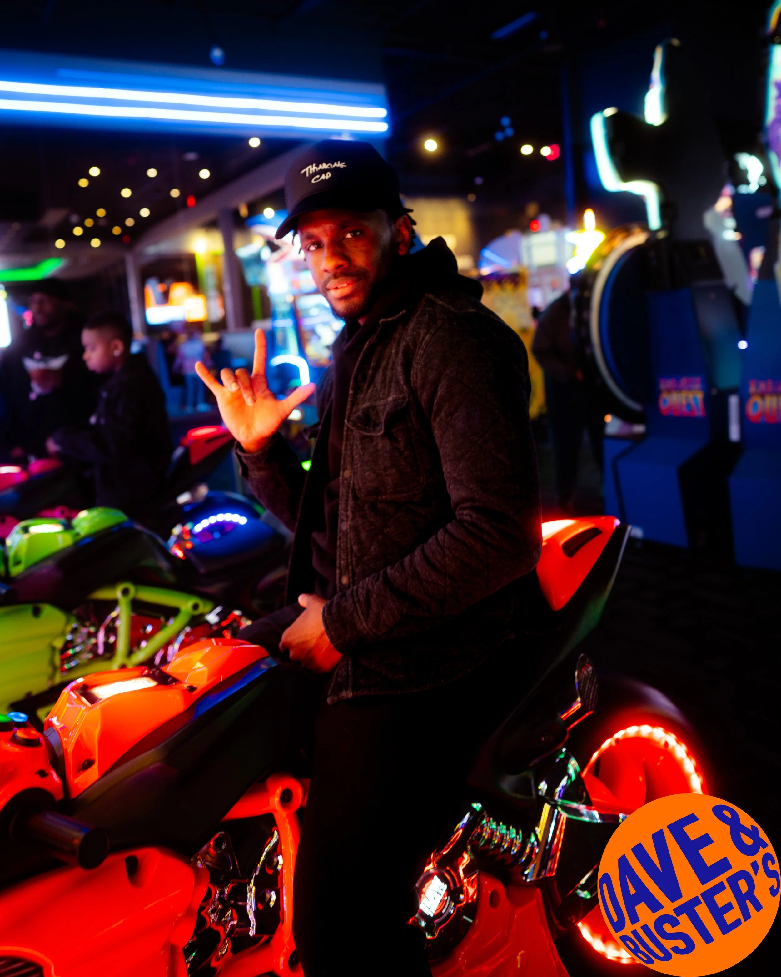 A man making a hand sign while riding an orange arcade motorcycle game in a dark arcade with colorful lights and other arcade machines in the background.