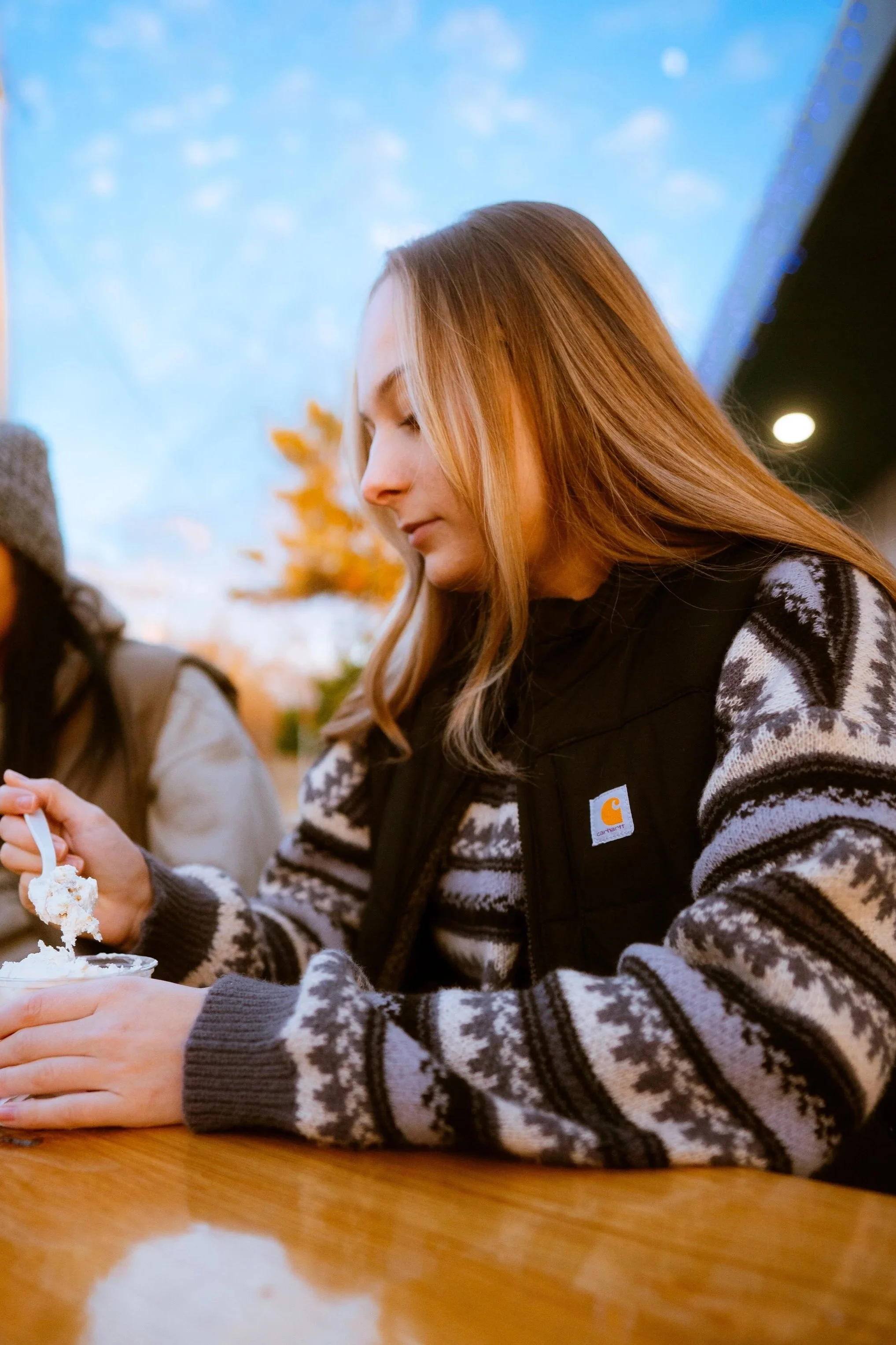 A woman with long red hair and a patterned sweater is sitting at a table, holding a spoonful of ice cream.