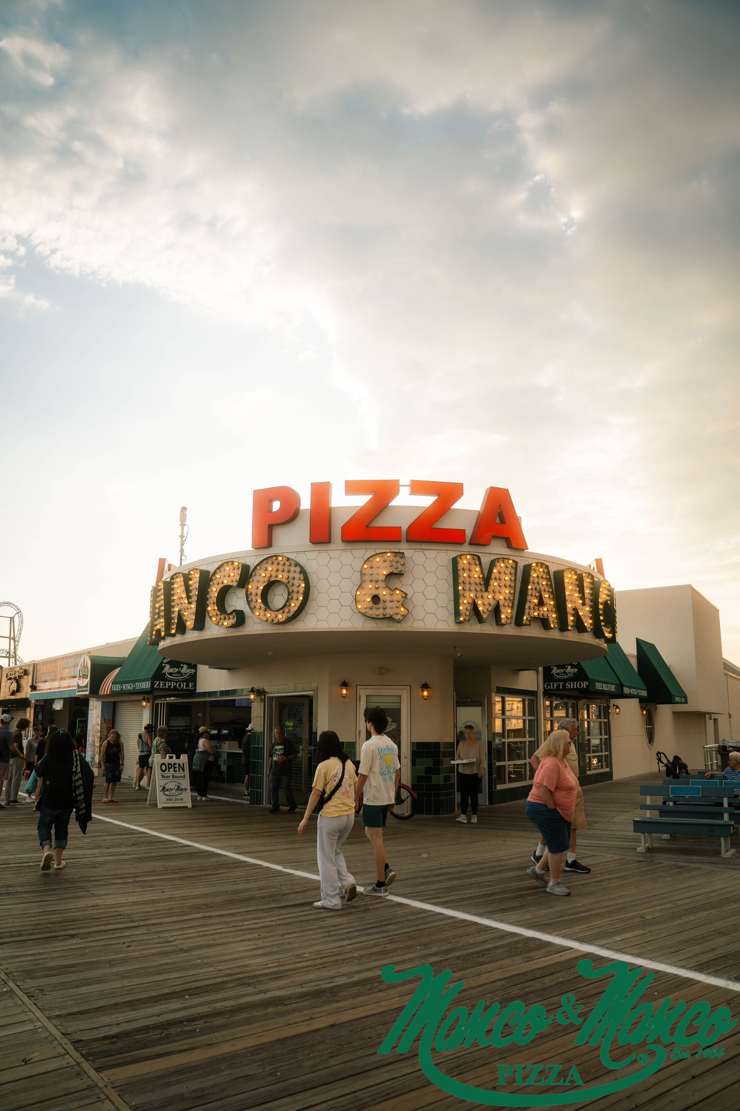 People walking on a wooden pier outside a pizza restaurant with a marquee sign and a green canopy, under a partly cloudy sky.