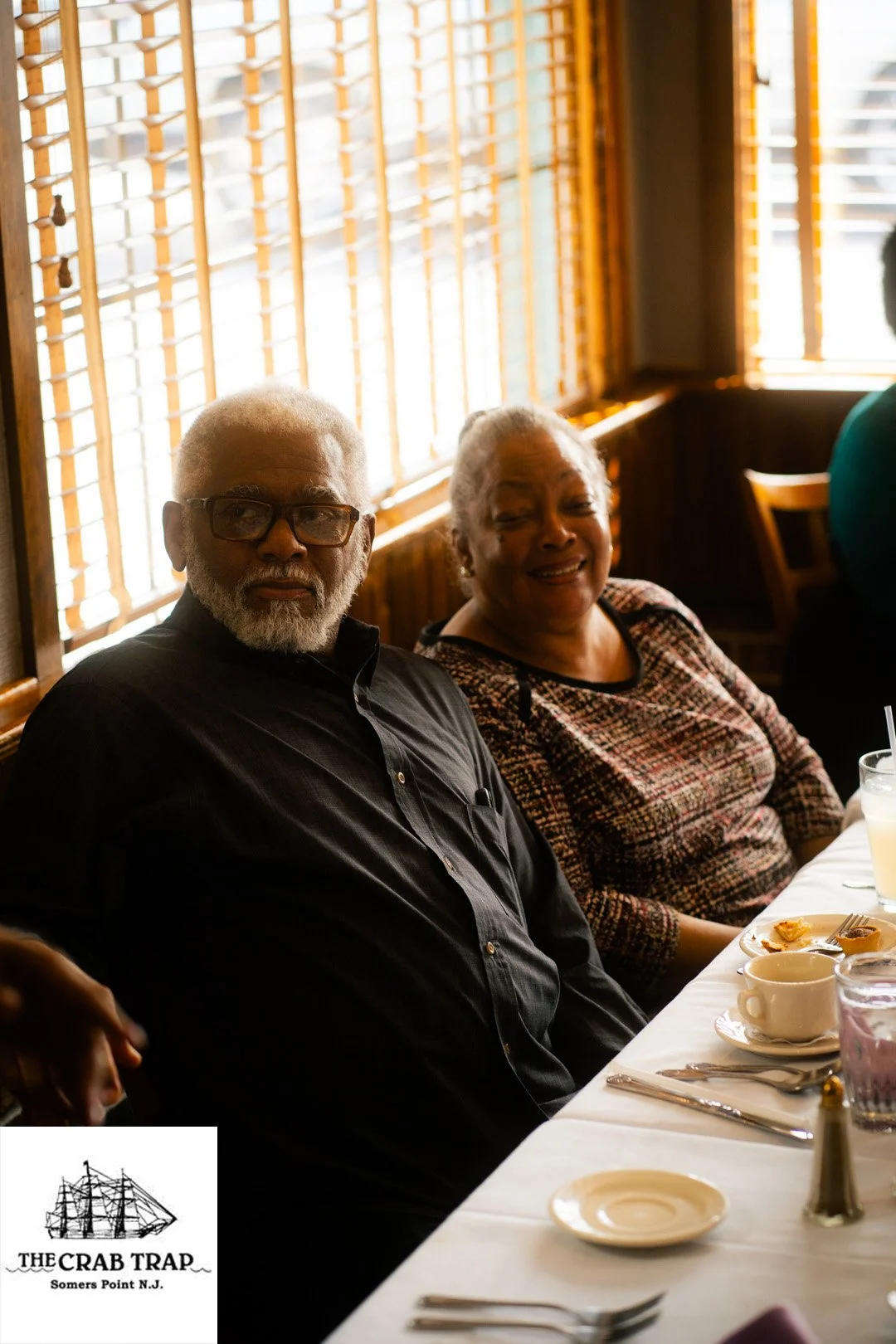 Two elderly people, a man with gray hair, beard, and glasses, and a woman with gray hair, sitting at a restaurant table with food and drinks, near a window with wooden blinds, in a warm lighting environment.