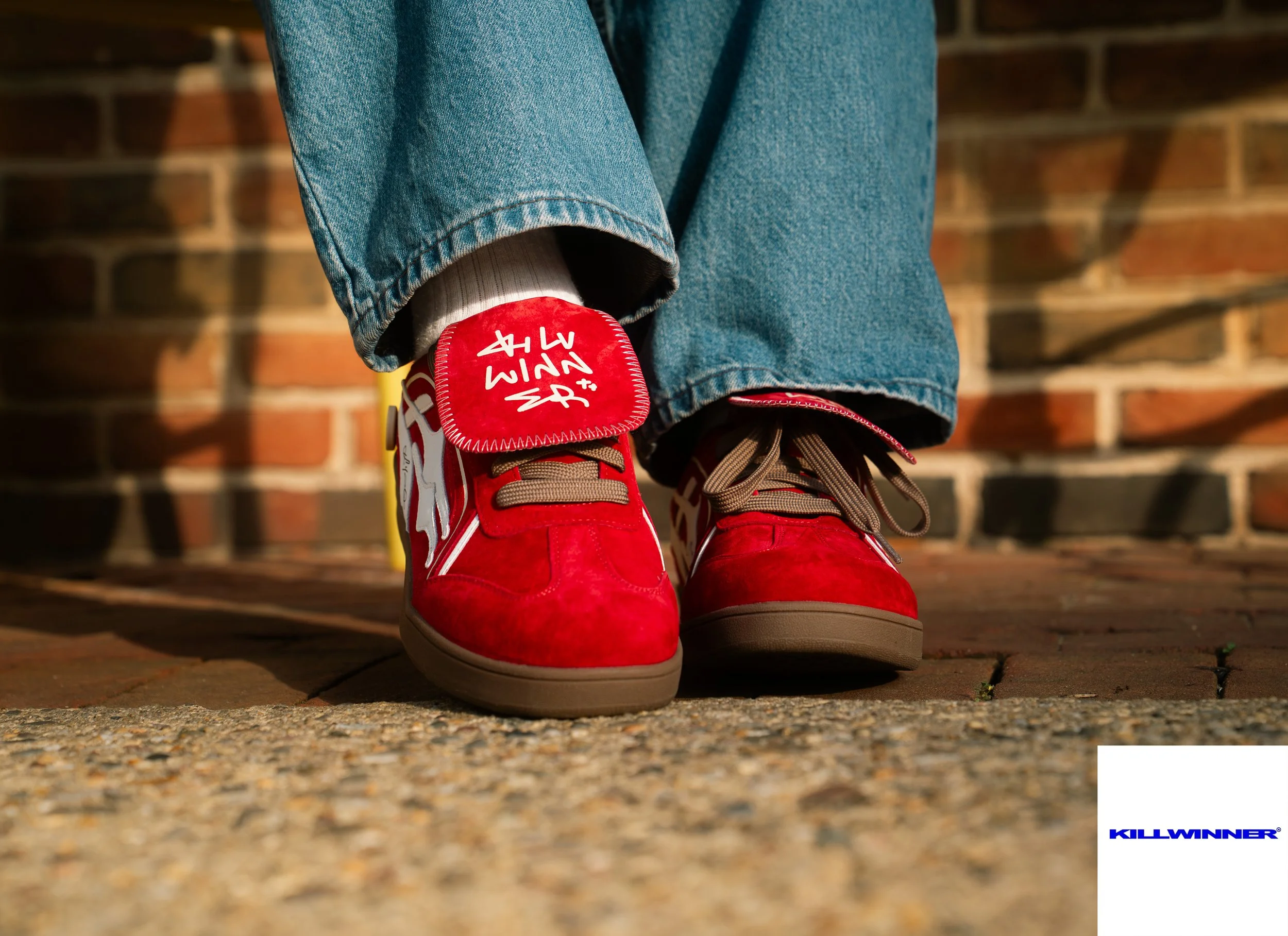 Close-up of a person's feet wearing red sneakers with brown laces and a logo tag, jeans, and white socks, standing on a paved road with a brick wall in the background.