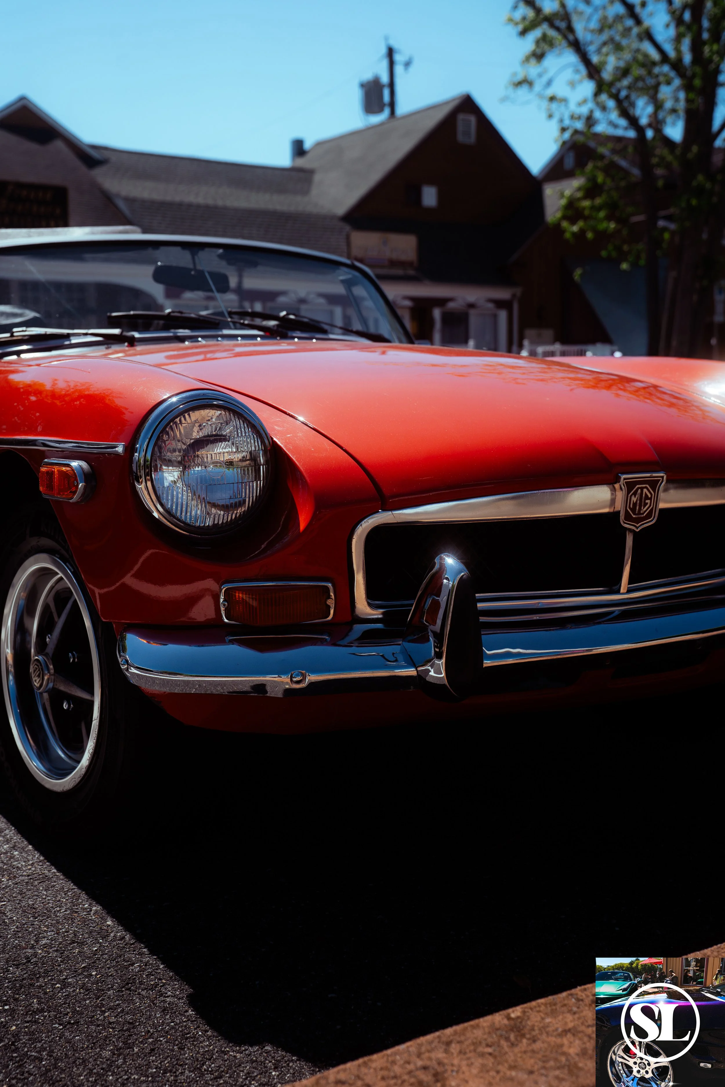 Front view of a red vintage MG convertible car parked outdoors on an asphalt surface, with houses, trees, and a utility pole in the background, under a clear blue sky.