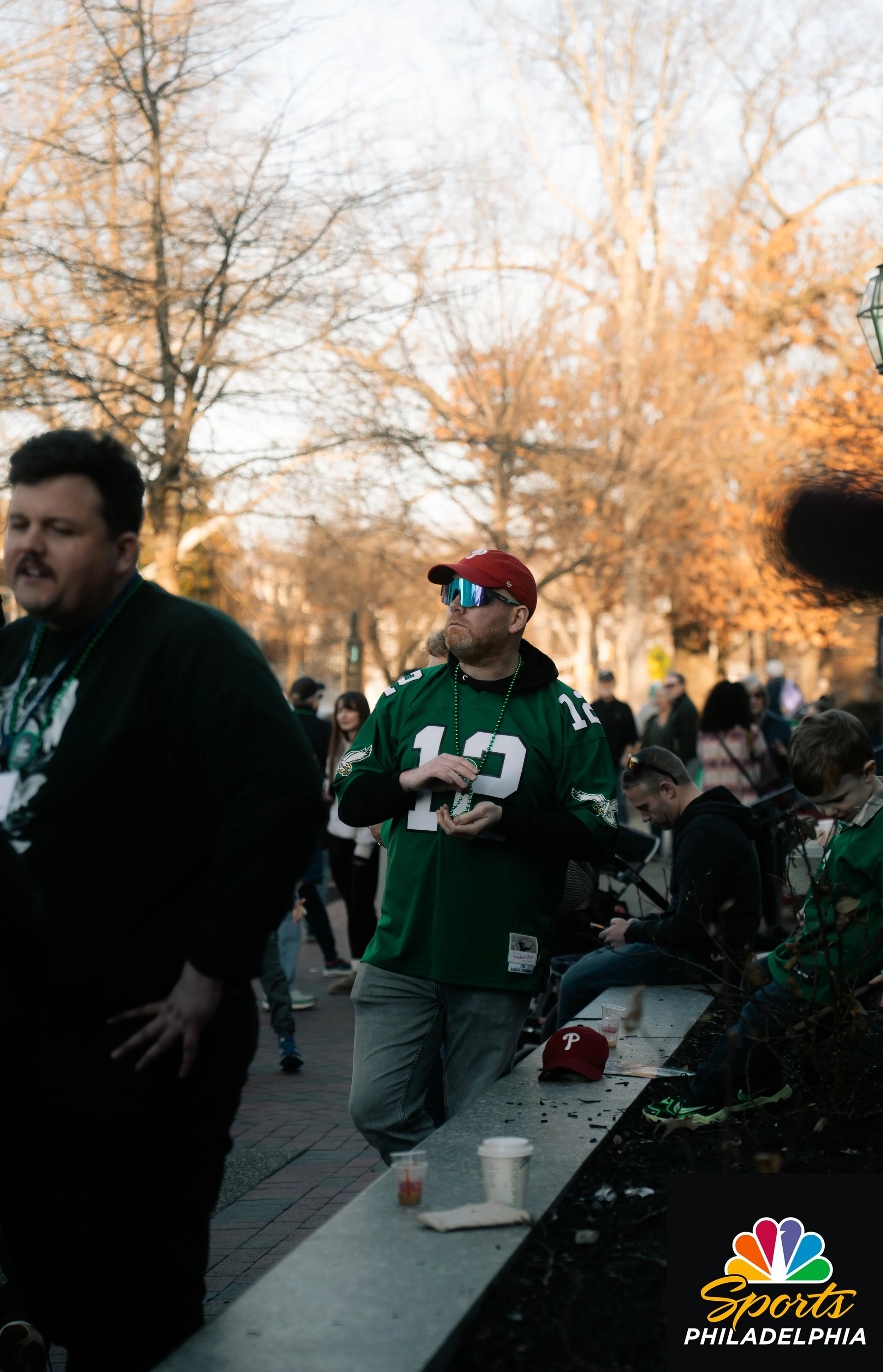 A group of people gathered outdoors in a park during fall, with a man wearing a Philadelphia Eagles jersey and sunglasses, standing near a bench with a Philadelphia Phillies cap on it, and NBC Sports Philadelphia logo in the corner.