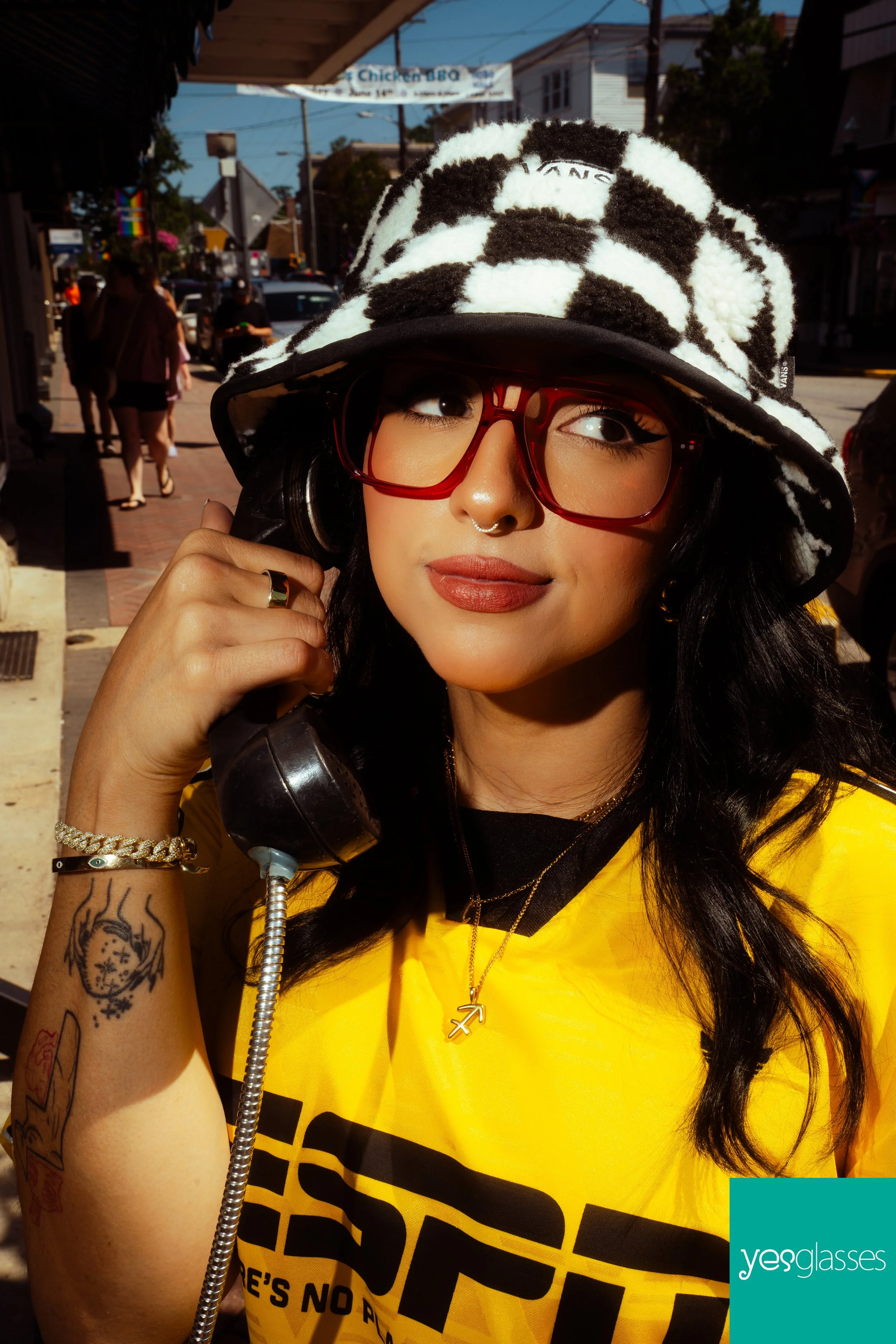 A young woman with black hair and tan skin wearing red glasses, a black and white fuzzy striped bucket hat, and a yellow T-shirt is holding a vintage black telephone receiver to her ear on a sunny street. She has tattoos on her left forearm and is we