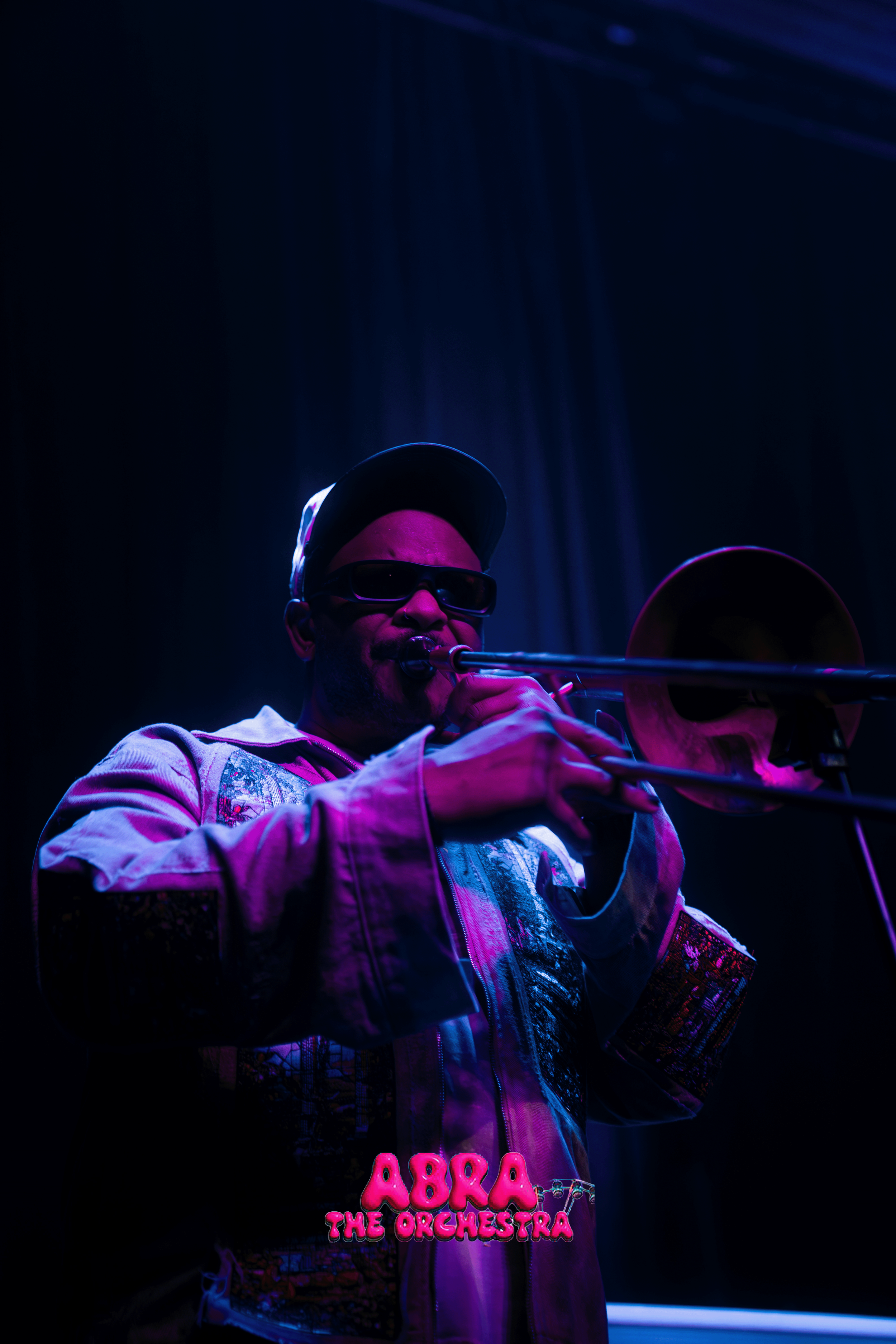 A musician wearing sunglasses, a cap, and a colorful jacket playing the trumpet on stage with a neon pink and green sign that reads 'Aabra and The Orchestra' in the background, illuminated with purple and blue lighting.