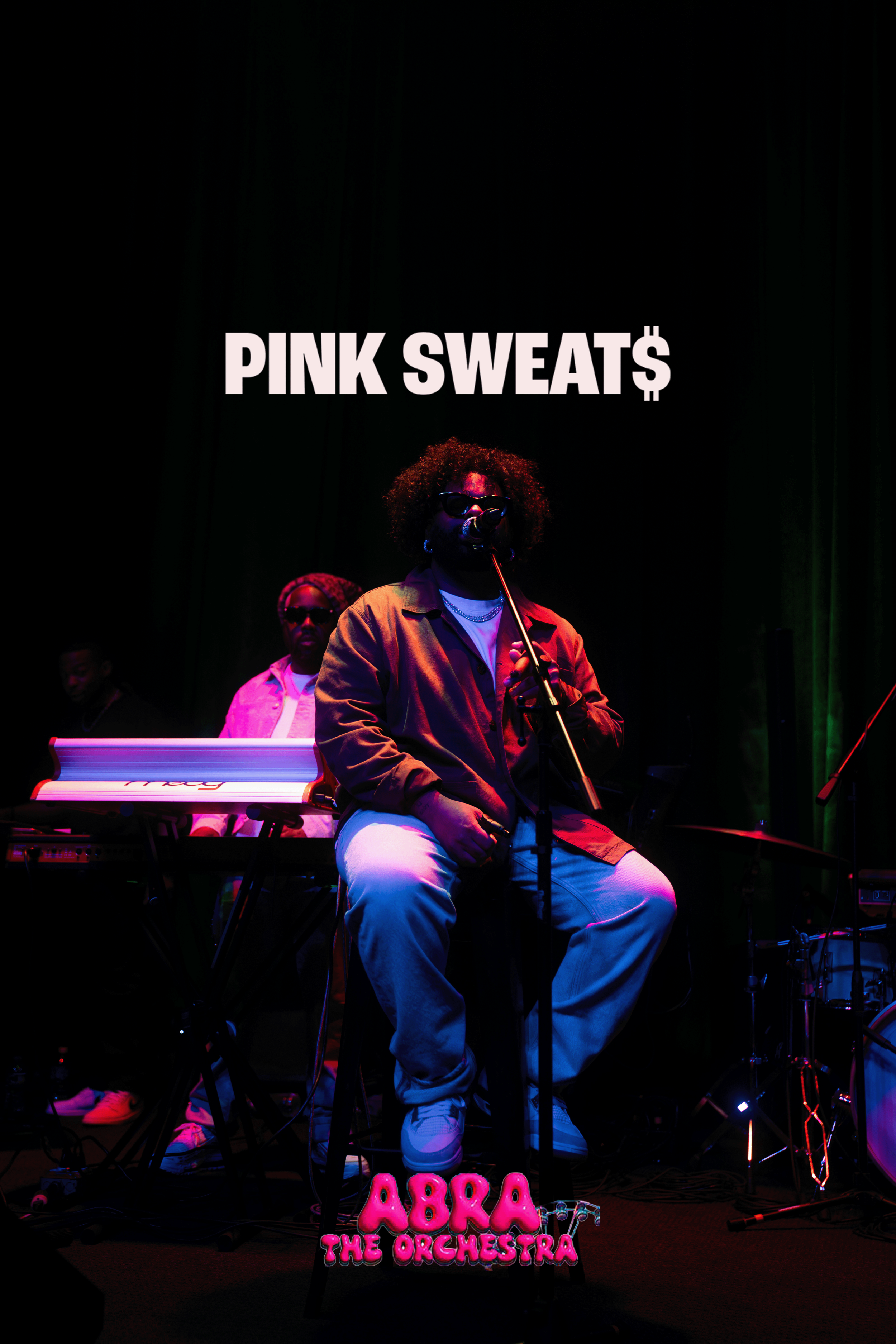 A woman singing into a microphone on stage with musicians in the background, illuminated by colorful stage lights. There is a sign with pink neon lights at the bottom saying "ABRA THE ORCHESTRA."