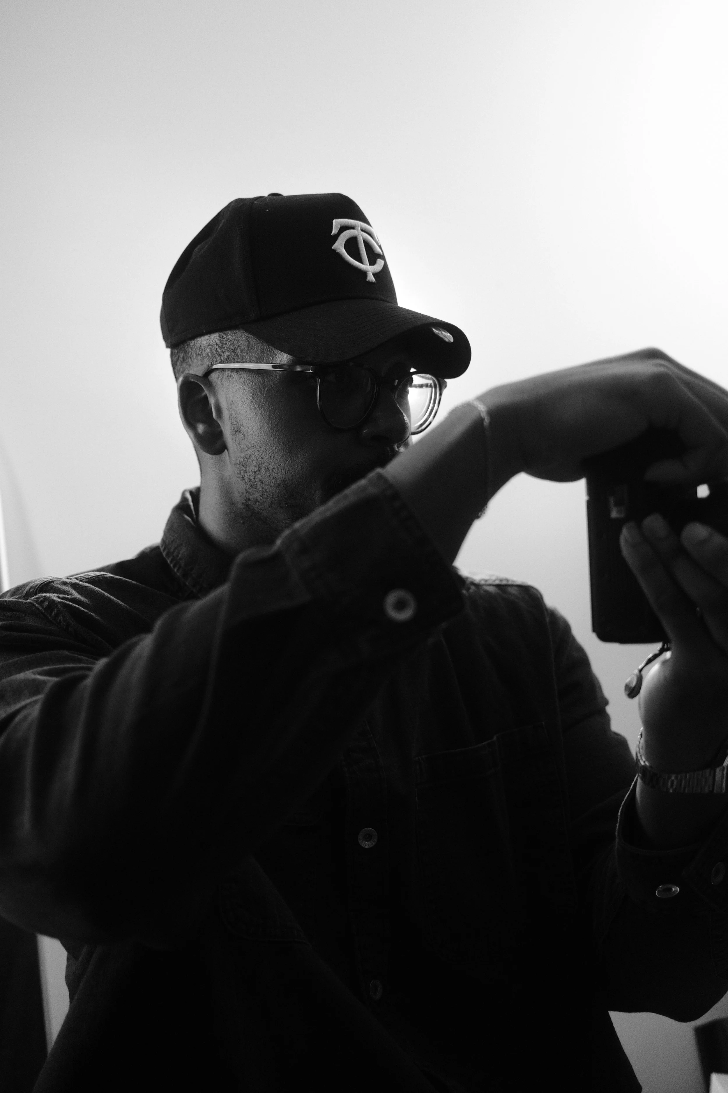 A person with short hair wearing glasses, a baseball cap with a logo, and a jacket taking a photo with a smartphone. The background is plain and light-colored.