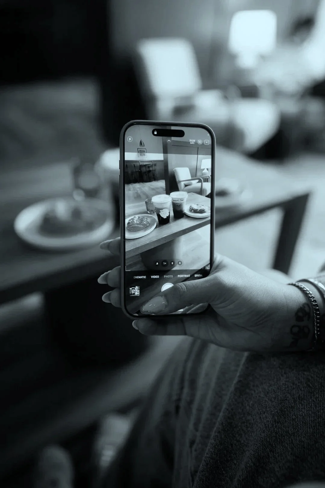 A person with a tattoo on their wrist holding a smartphone, photographing a table of food including drinks and pancakes, in a café.